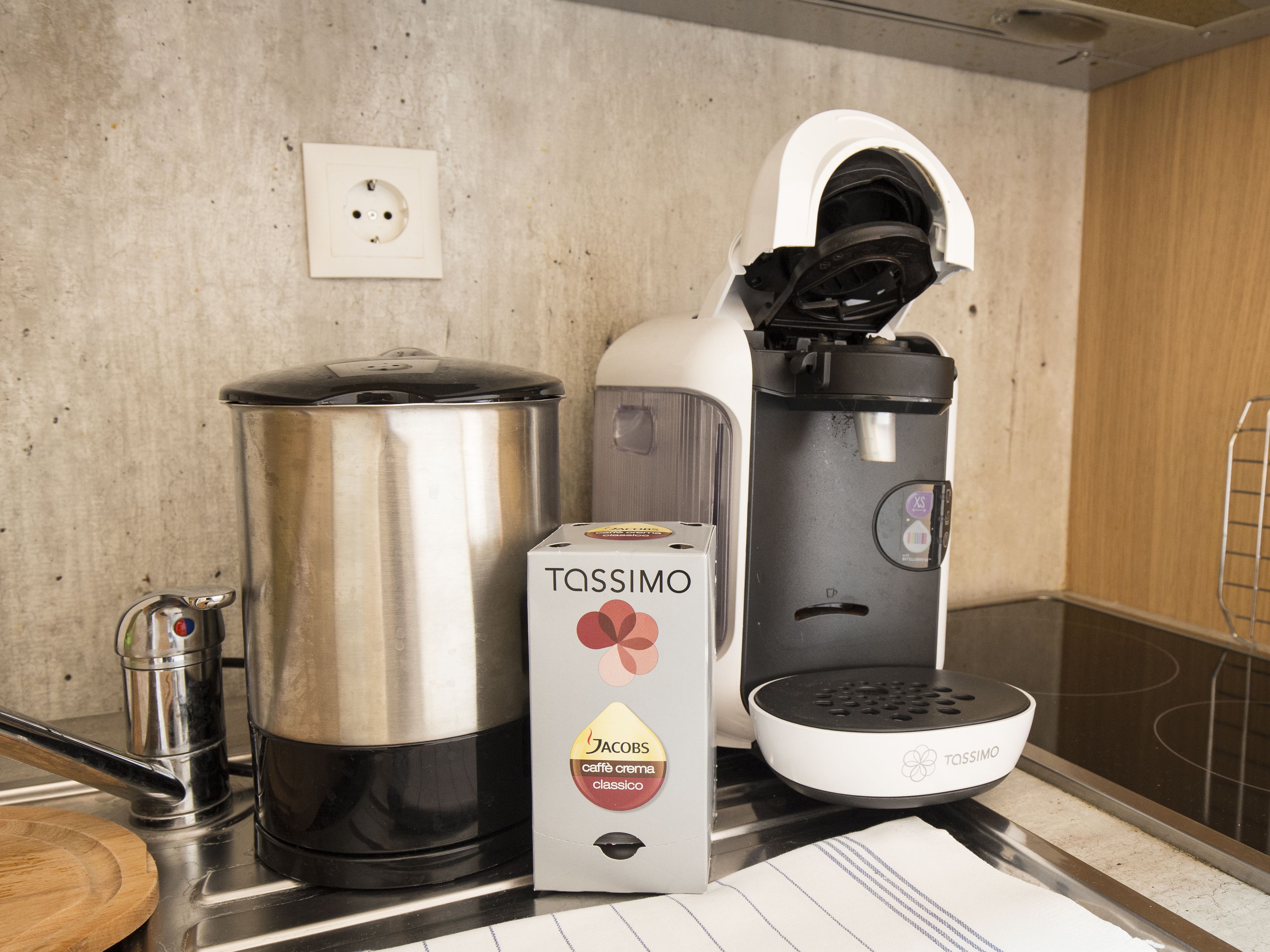 Una cocina con cafetera, hervidor, una olla grande y una caja de café con la etiqueta 'Tassimo'.