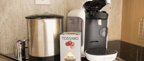 Una cocina con cafetera, hervidor, una olla grande y una caja de café con la etiqueta 'Tassimo'.