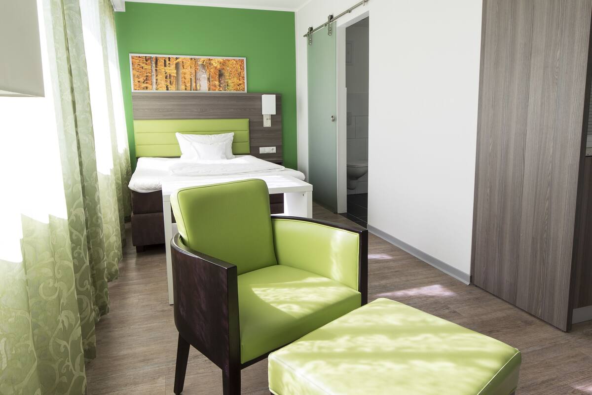 Habitación de hotel con cama, una silla, un banco, un cuadro en la pared y una puerta corrediza.