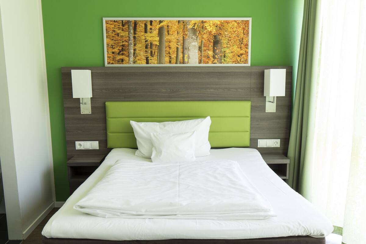 Una cama bien hecha con ropa blanca, un cabecero tapizado en verde y un cuadro con árboles otoñales encima.