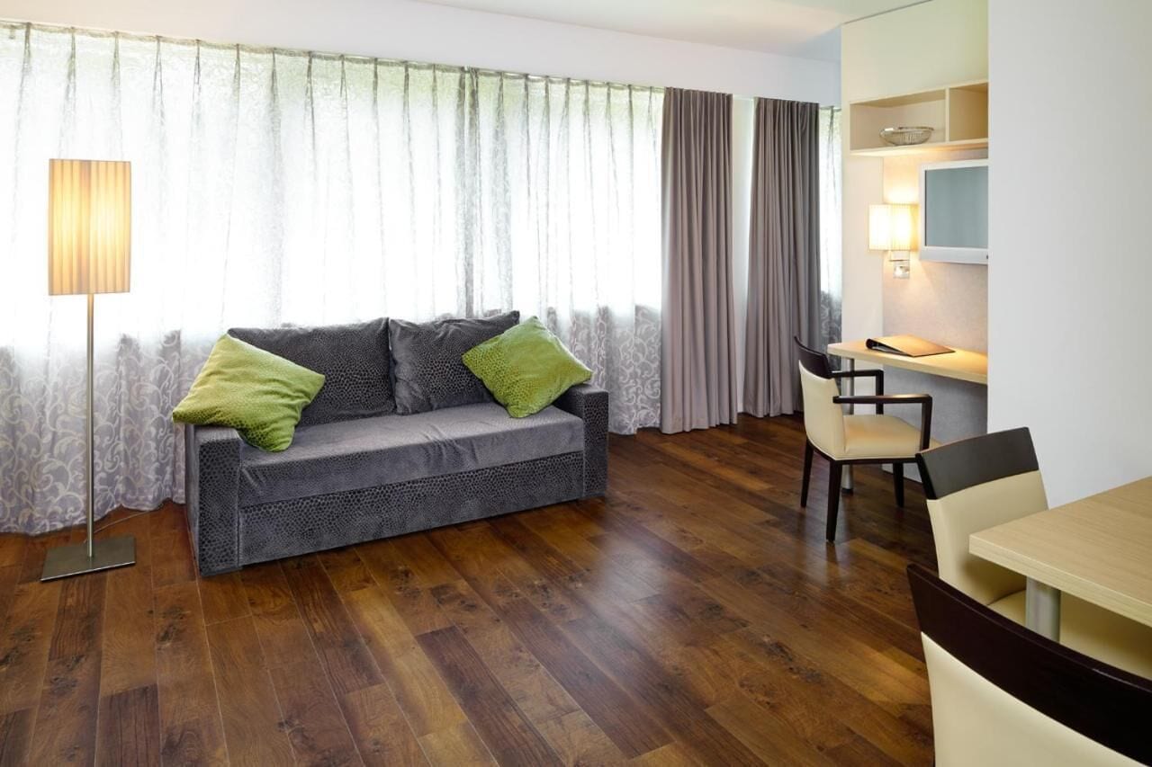A modern living room with a grey sofa, a wooden floor, a dining table with chairs, and a TV on the wall.