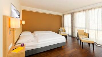 A hotel room with a large bed, a desk with a lamp, a chair, and a window with curtains.