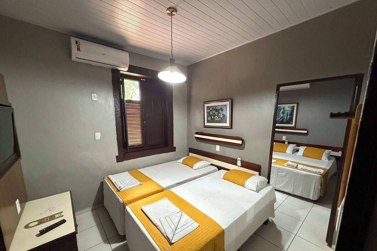 Une chambre d’hôtel avec deux lits, un bureau avec une chaise, un climatiseur, une fenêtre avec des stores et un ventilateur de plafond.