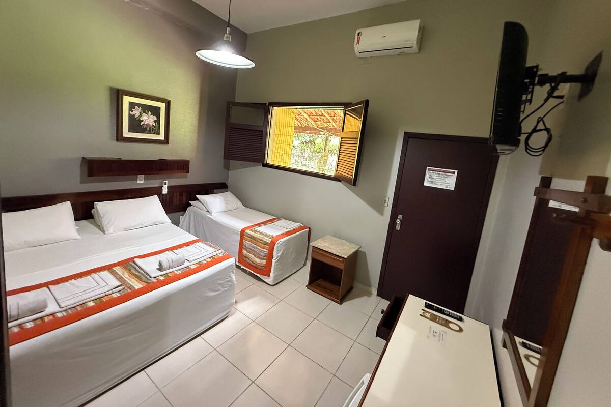 Une chambre d’hôtel avec deux lits, une petite table, une armoire, un appareil de climatisation, un luminaire au plafond et une fenêtre avec des stores.
