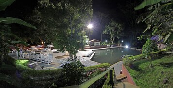Vista nocturna de una zona de piscina con un edificio pequeño, rodeada de vegetación exuberante y mobiliario exterior blanco.