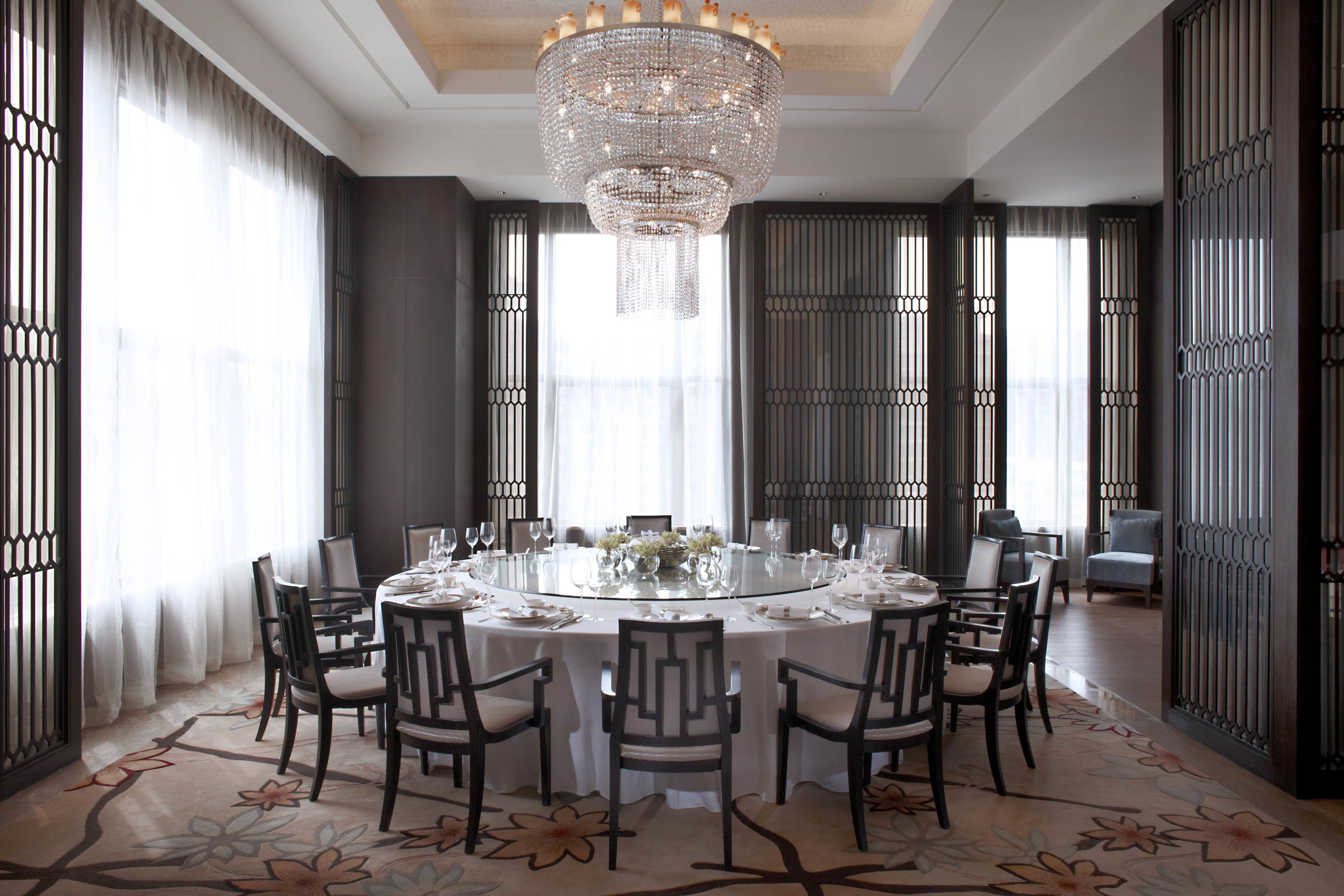 Un comedor formal con una mesa redonda dispuesta para ocho personas, rodeada de sillas, una araña y ventanas grandes con cortinas.