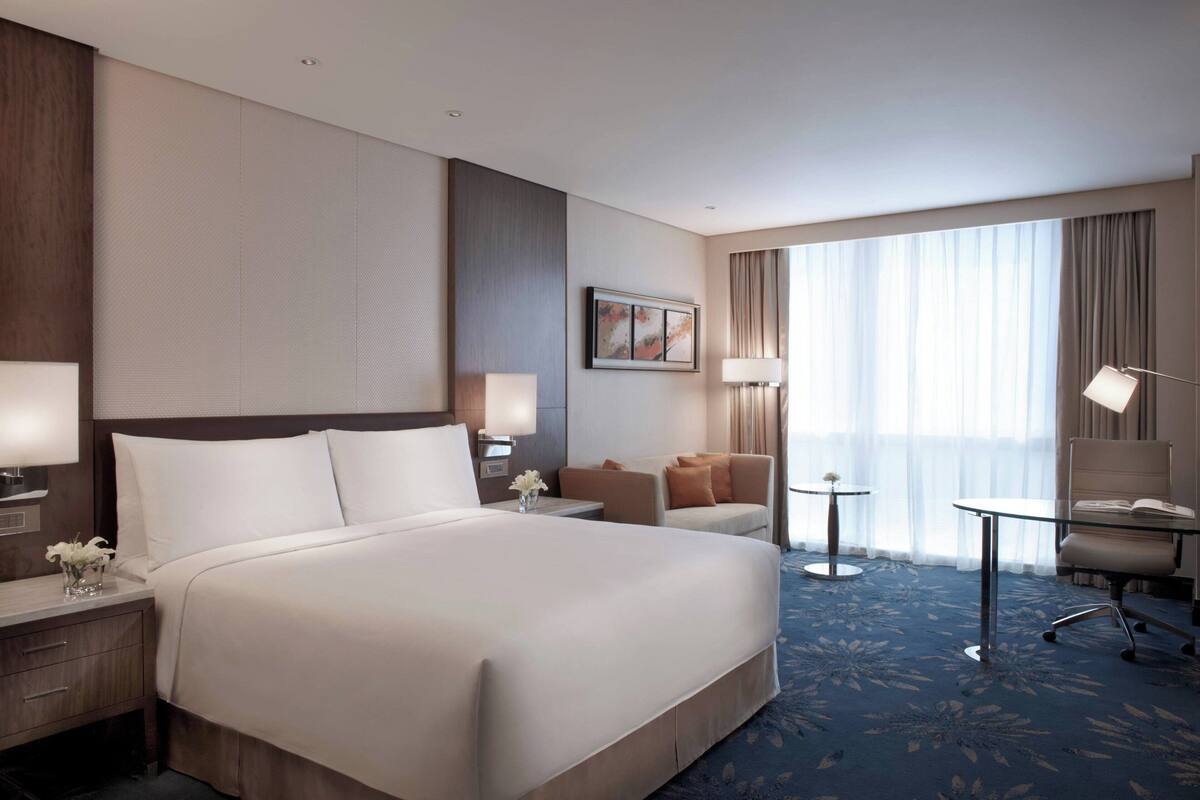 Een hotelkamer met een groot bed, een bureau met lamp, een stoel, een klein tafeltje en een raam met gordijnen.