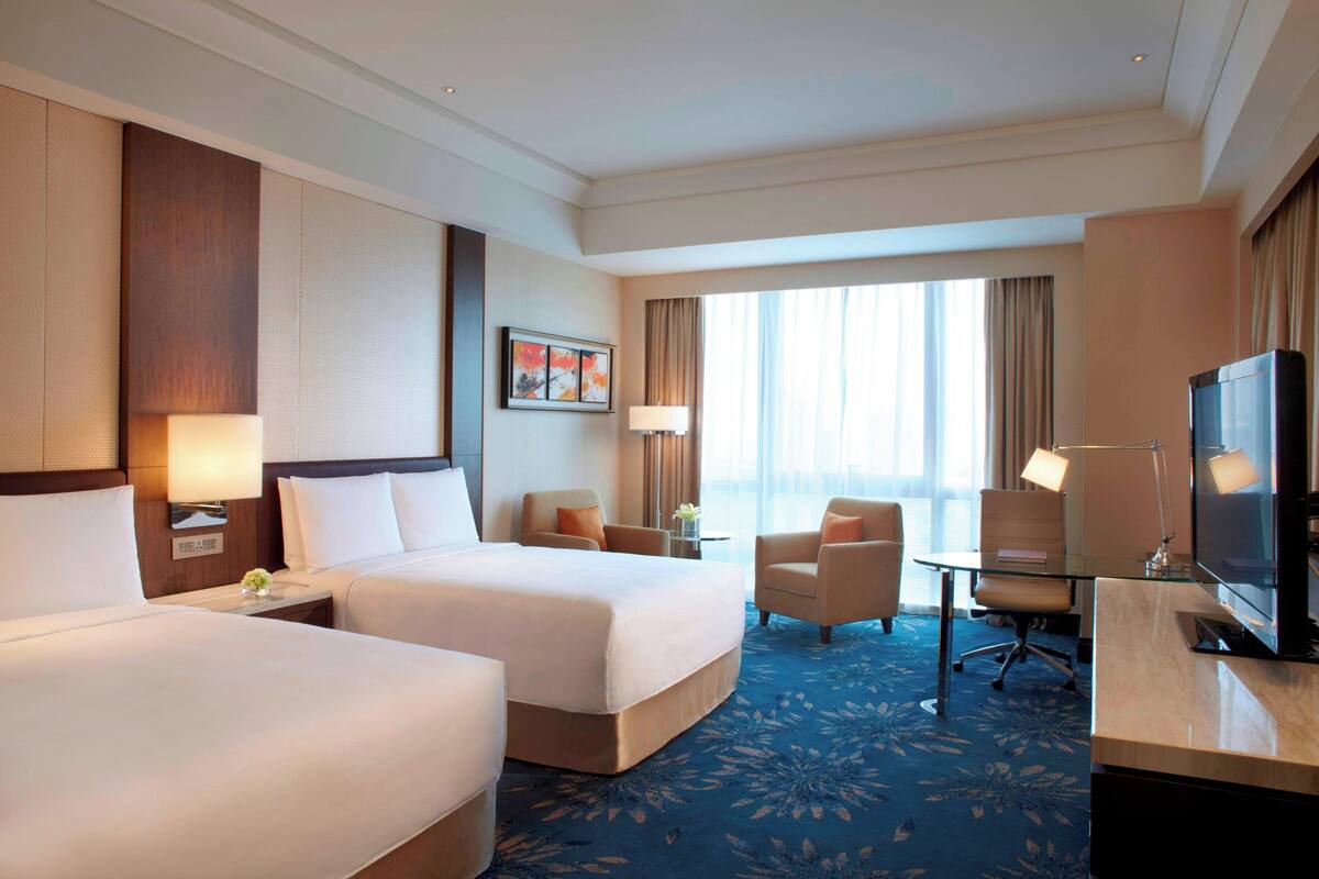 Een hotelkamer met twee bedden, een bureau met een televisie, een stoel, een lamp en een raam met gordijnen.