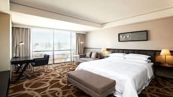A hotel room with a large bed, a desk, a chair, a bench, and a view of the city through large windows.