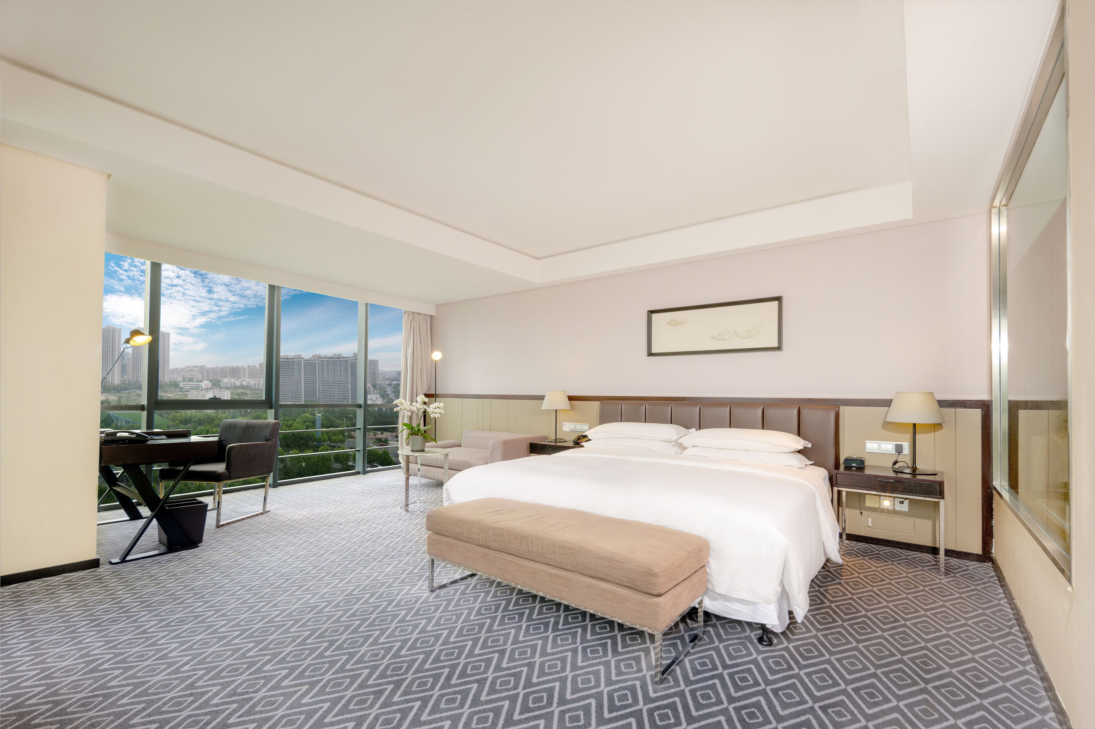 A hotel room with a large bed, a desk, and a chair. There is a window with a view of buildings and a lamp on the desk.