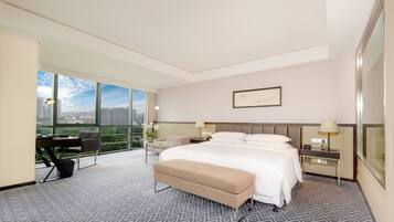 A hotel room with a large bed, a desk, and a chair. There is a window with a view of buildings and a lamp on the desk.