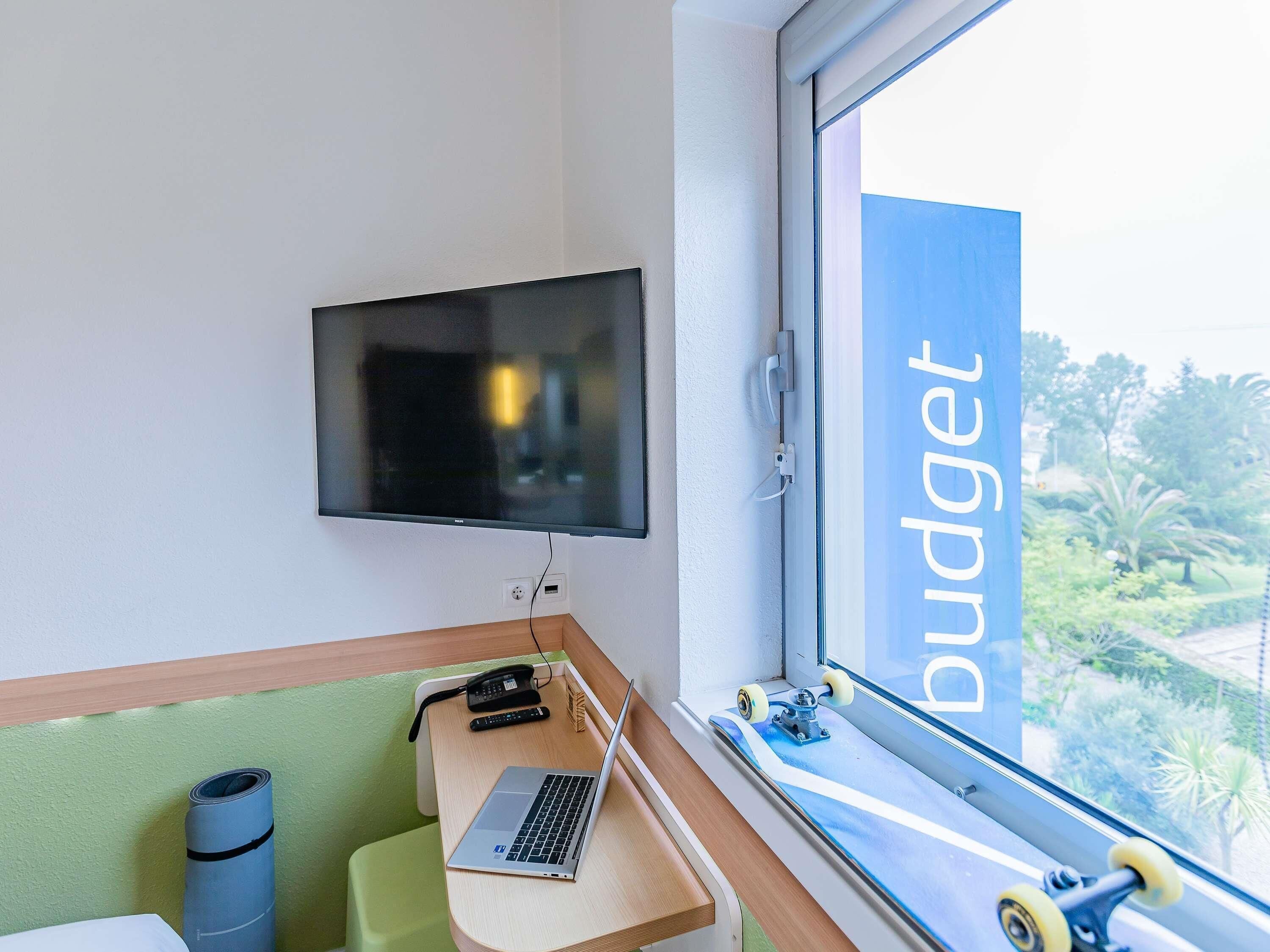A room with a TV mounted on the wall, a desk with a laptop, a phone, and a skateboard by the window.