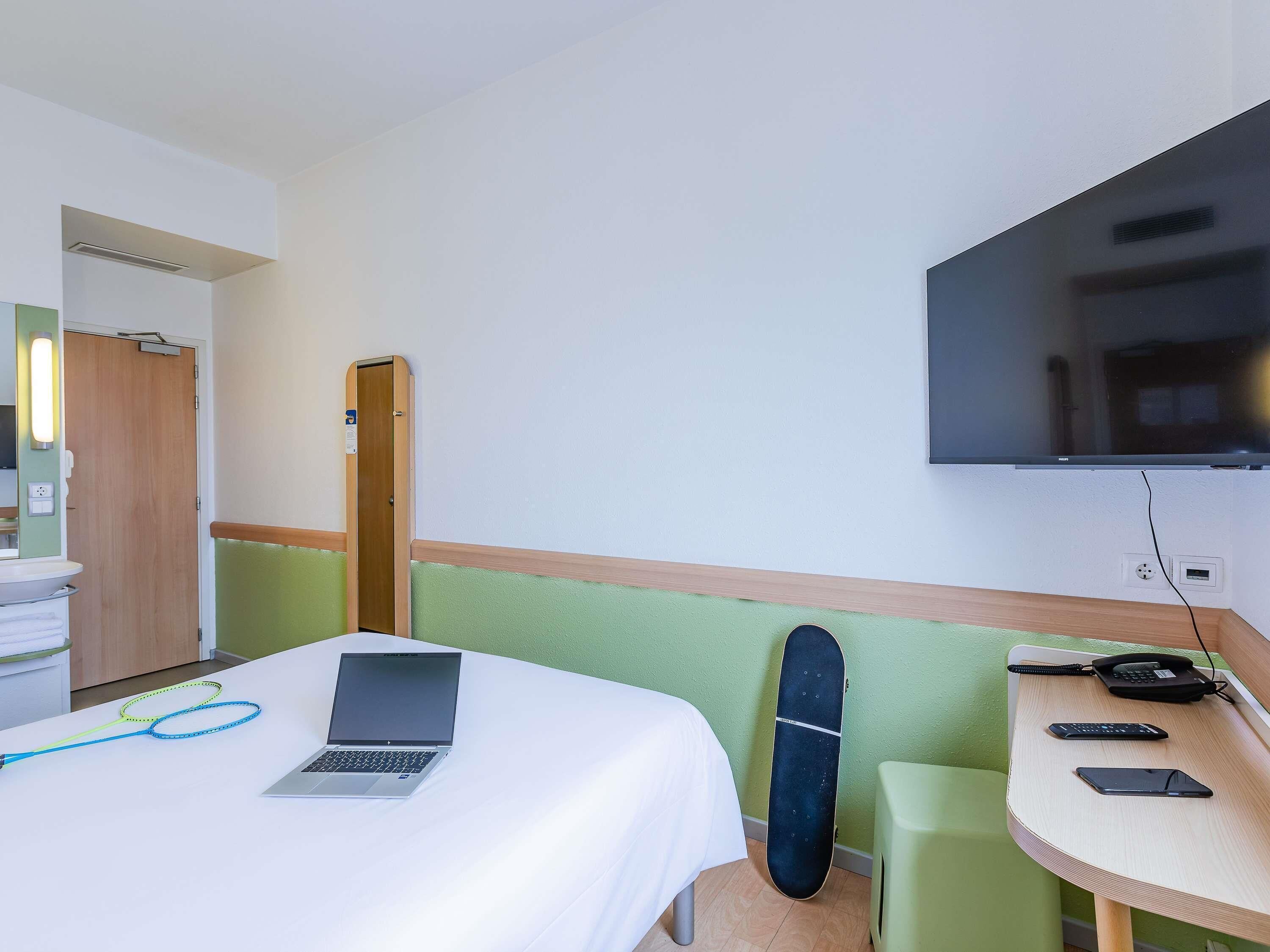 Una habitación de hotel con una cama, un escritorio con una computadora portátil, un monopatín, un televisor y un teléfono.