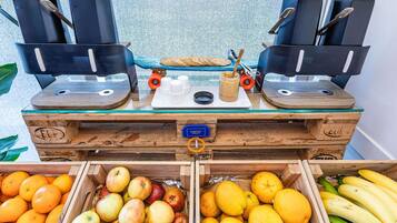 Een koffiehoek met espresso machines, een dienblad met kopjes en een drankje, en een houten kist met fruit zoals appels en sinaasappels.
