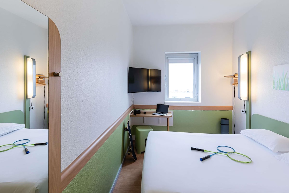 A hotel room with two beds, a desk with a laptop, a TV, and a window.