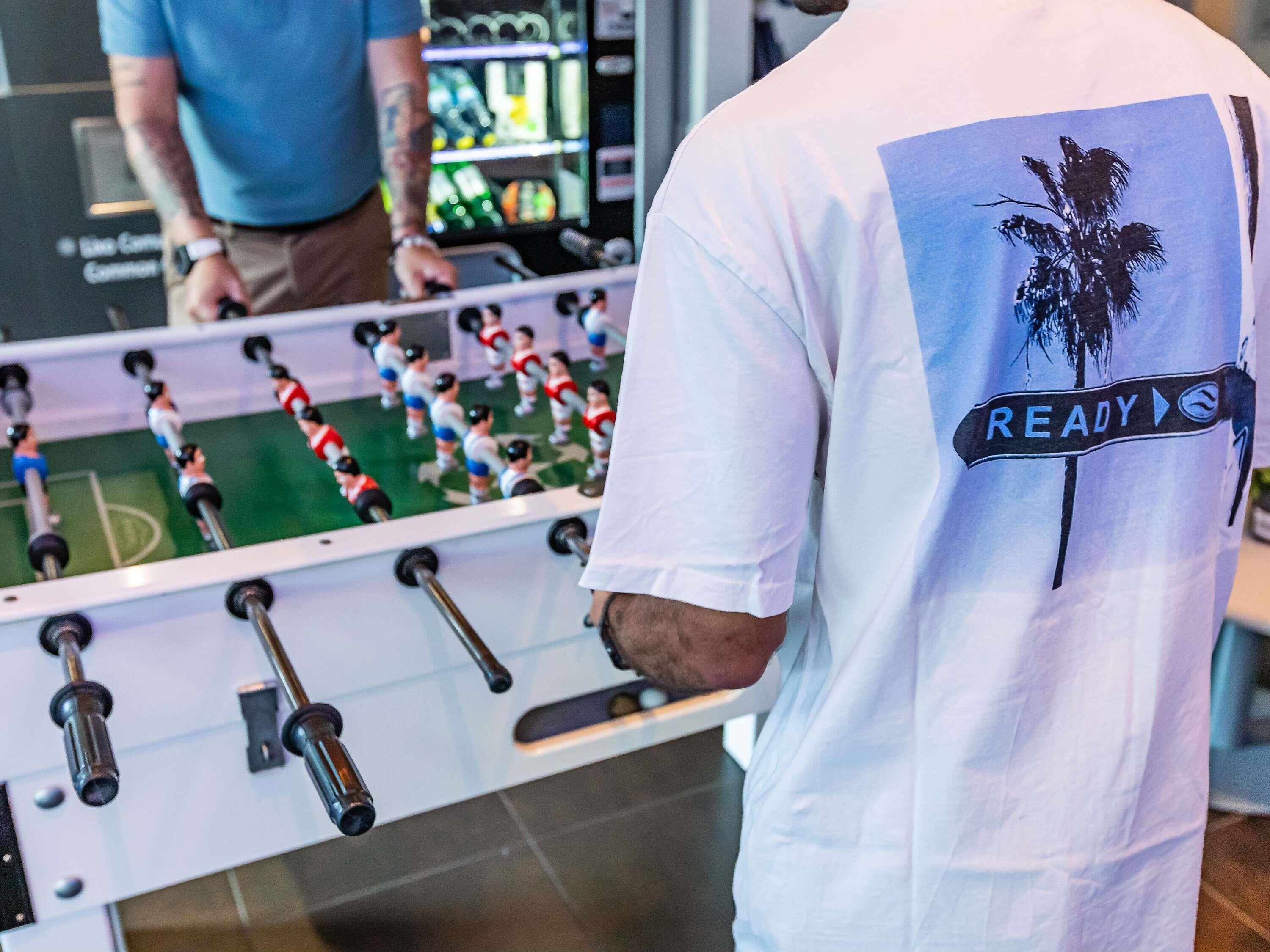 Una persona de pie frente a una mesa de fútbol de mesa, con un cartel que dice 'READY' en su camiseta.