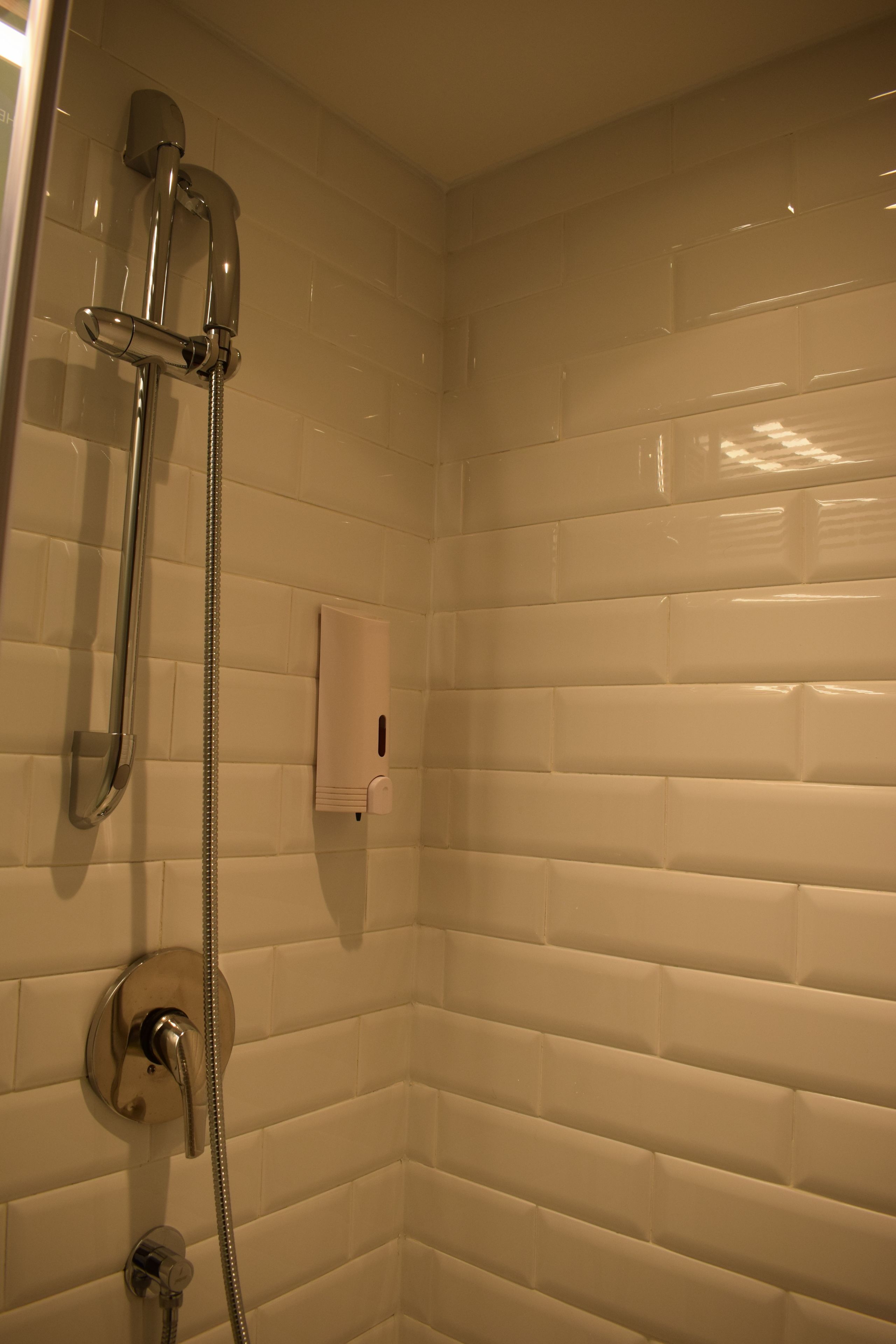 Une salle de bain avec des murs carrelés de carreaux blancs, une douche avec un pommeau de douche amovible et un distributeur de savon fixé au mur.