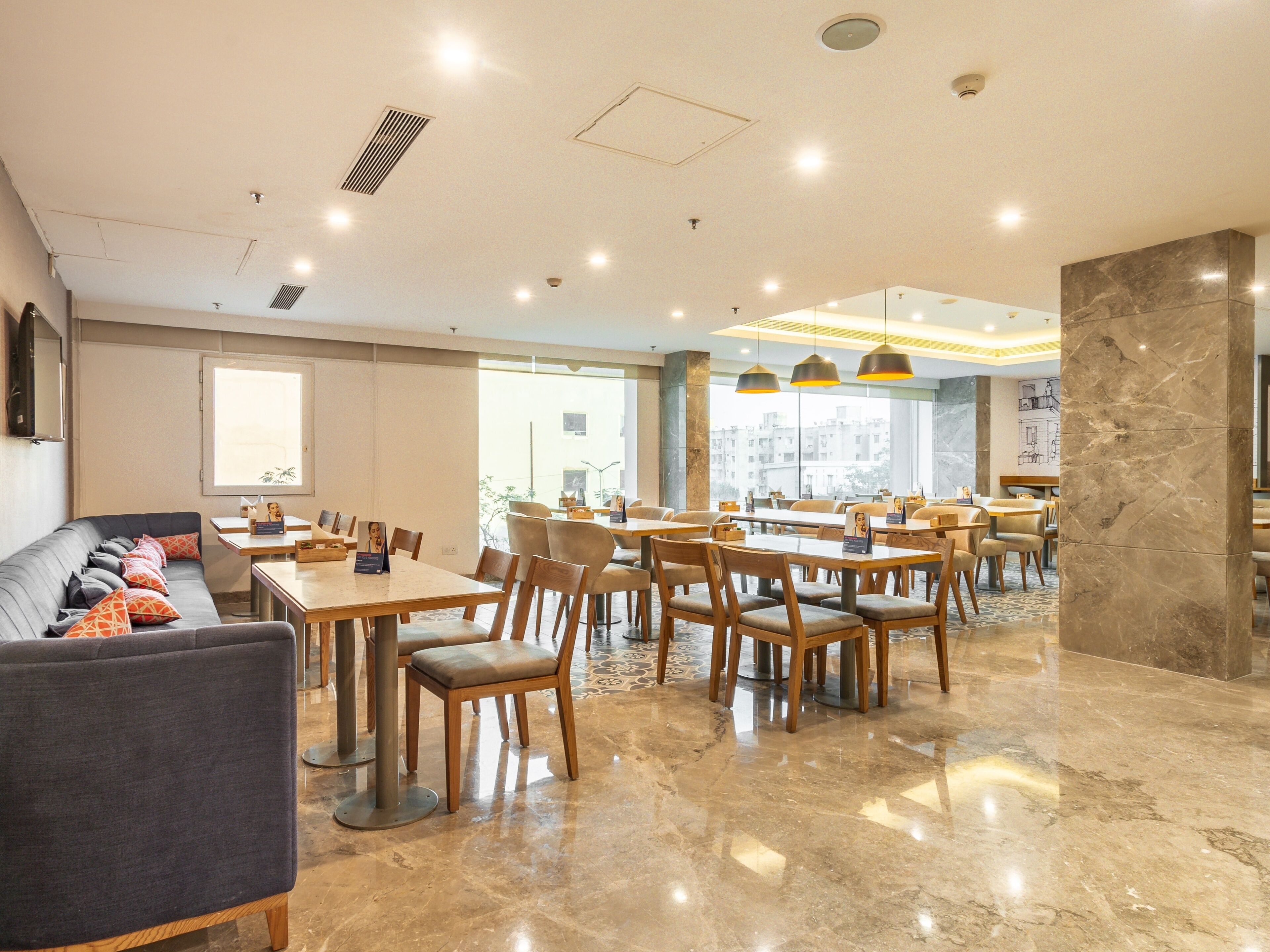A spacious dining area with multiple tables and chairs, a sofa, and a large window providing a view of the cityscape.