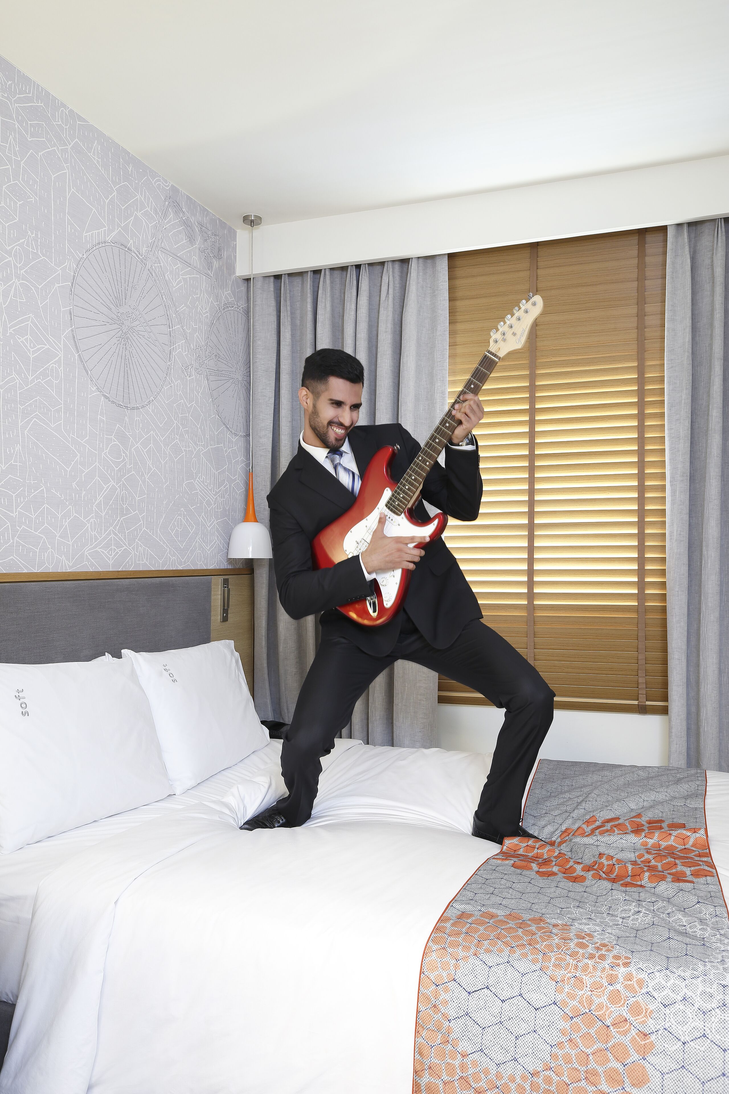A man in a suit playing an electric guitar on a bed in a hotel room.