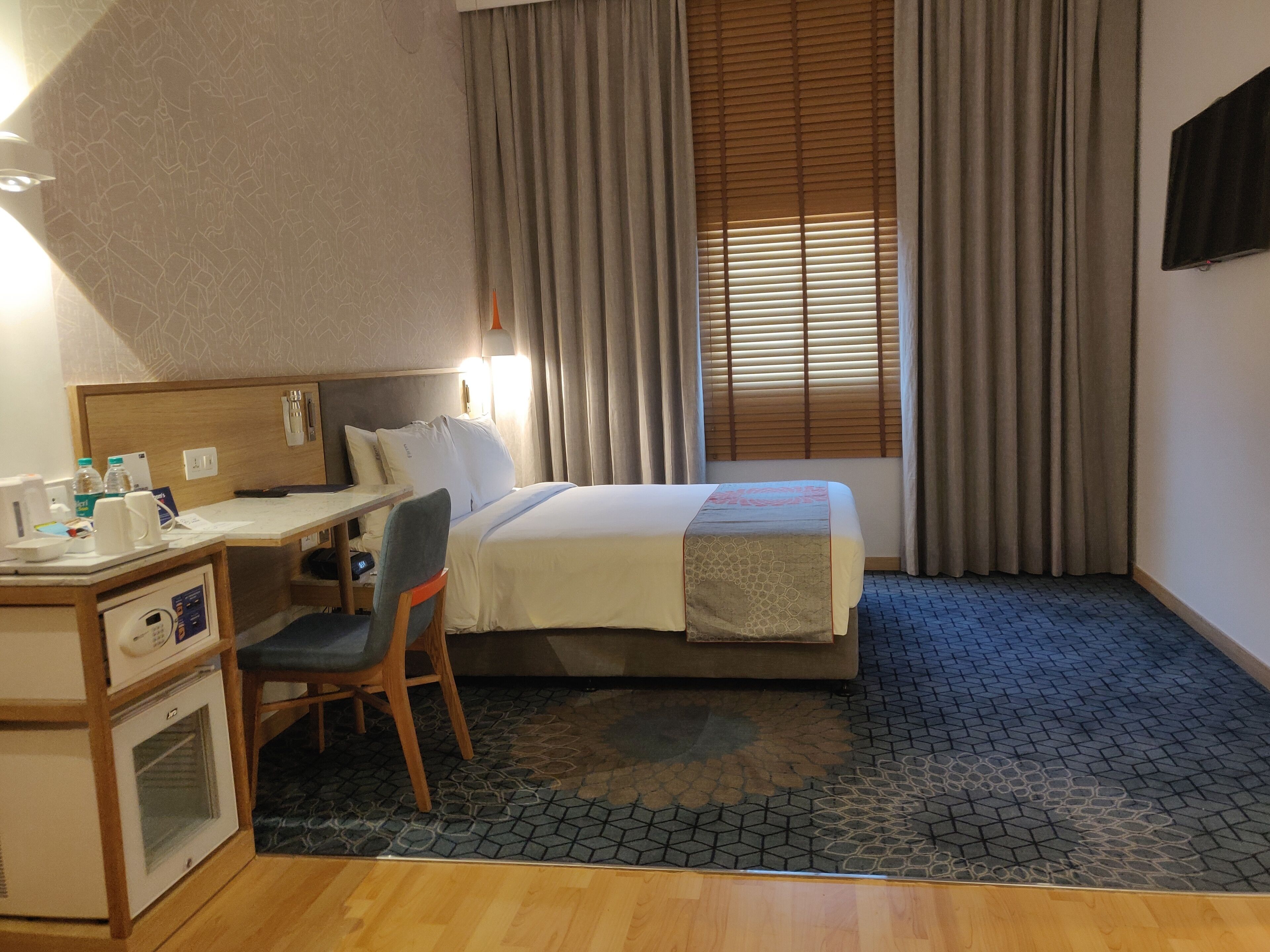 A hotel room with a bed, a desk, a chair, a small kitchenette, and a window with curtains.