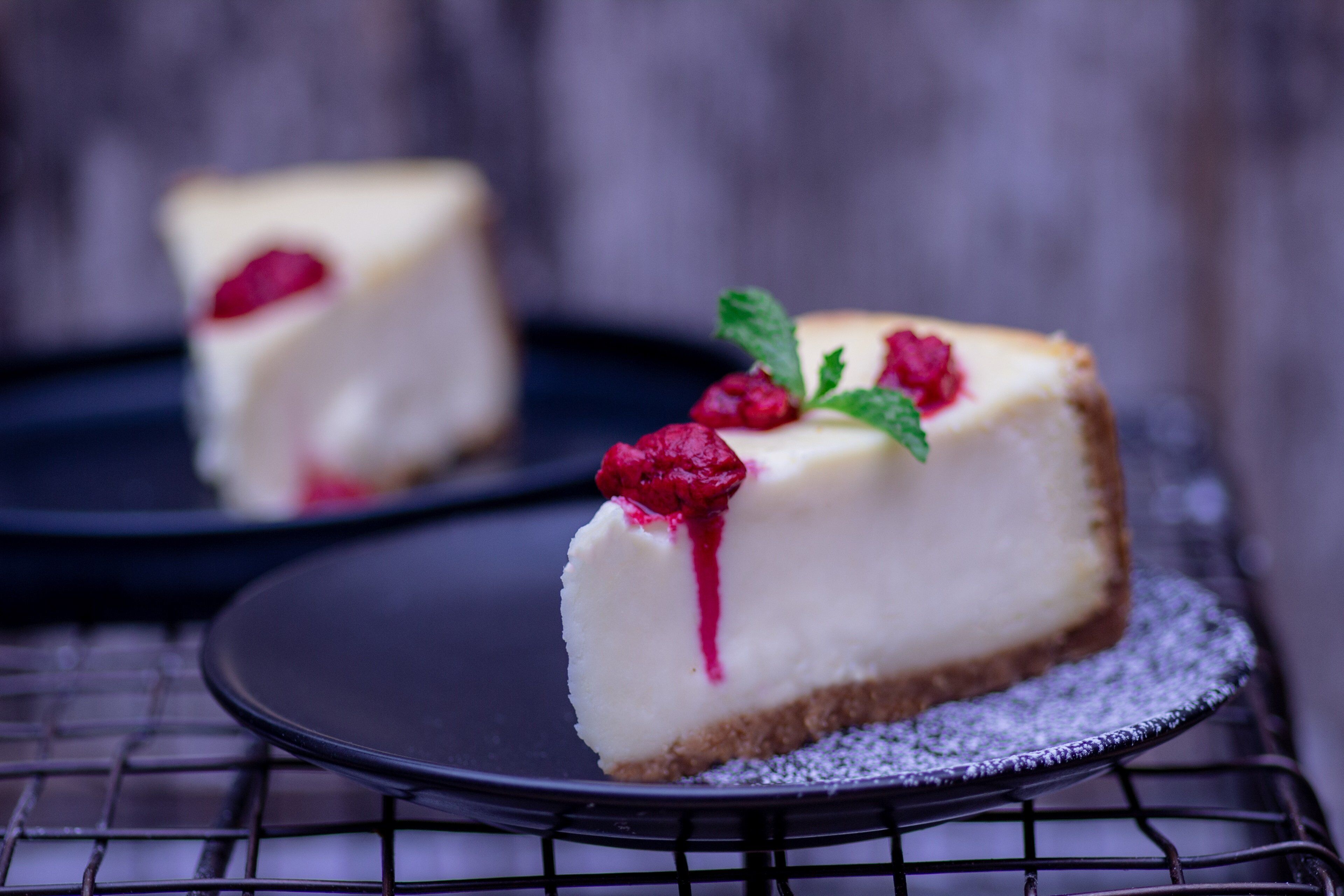 A slice of cheesecake on a black plate with a dusting of powdered sugar, garnished with a mint leaf and raspberries.