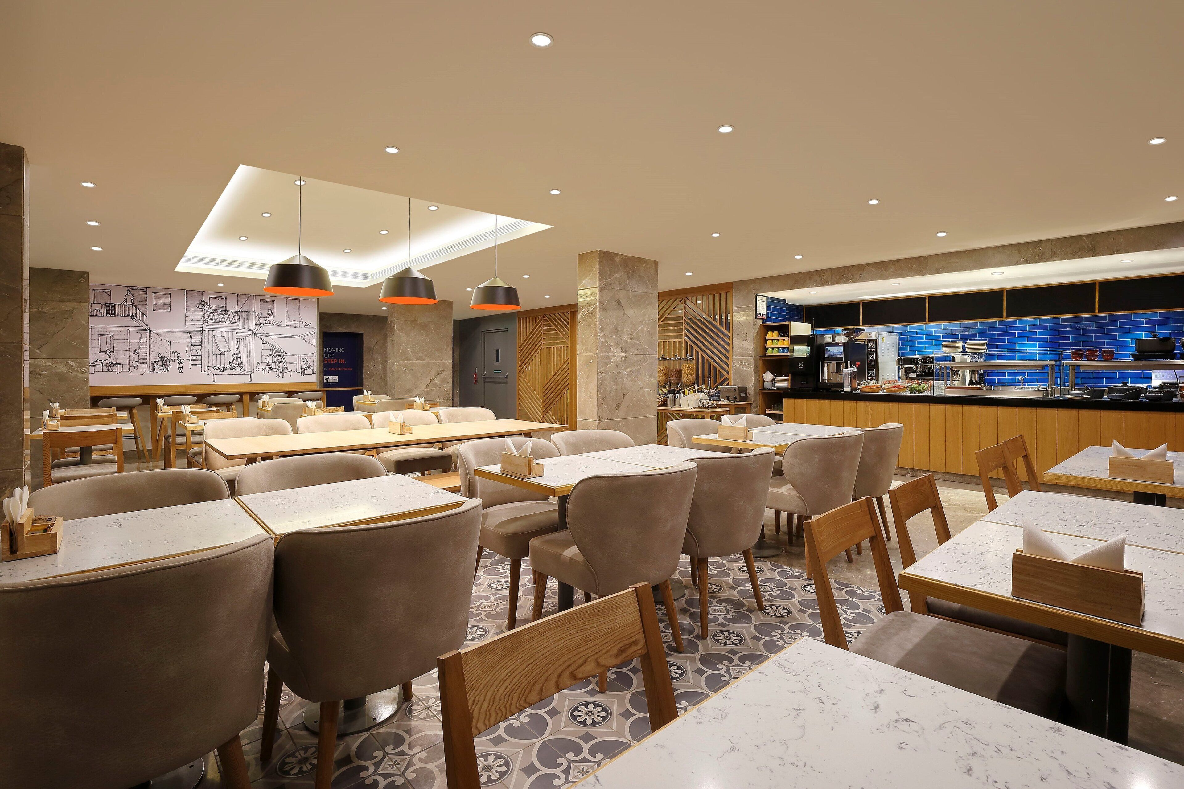 Un restaurant moderne doté d’un sol en marbre, de tables et de chaises en bois, d’un éclairage suspendu et d’un espace cuisine visible à travers une paroi en verre.