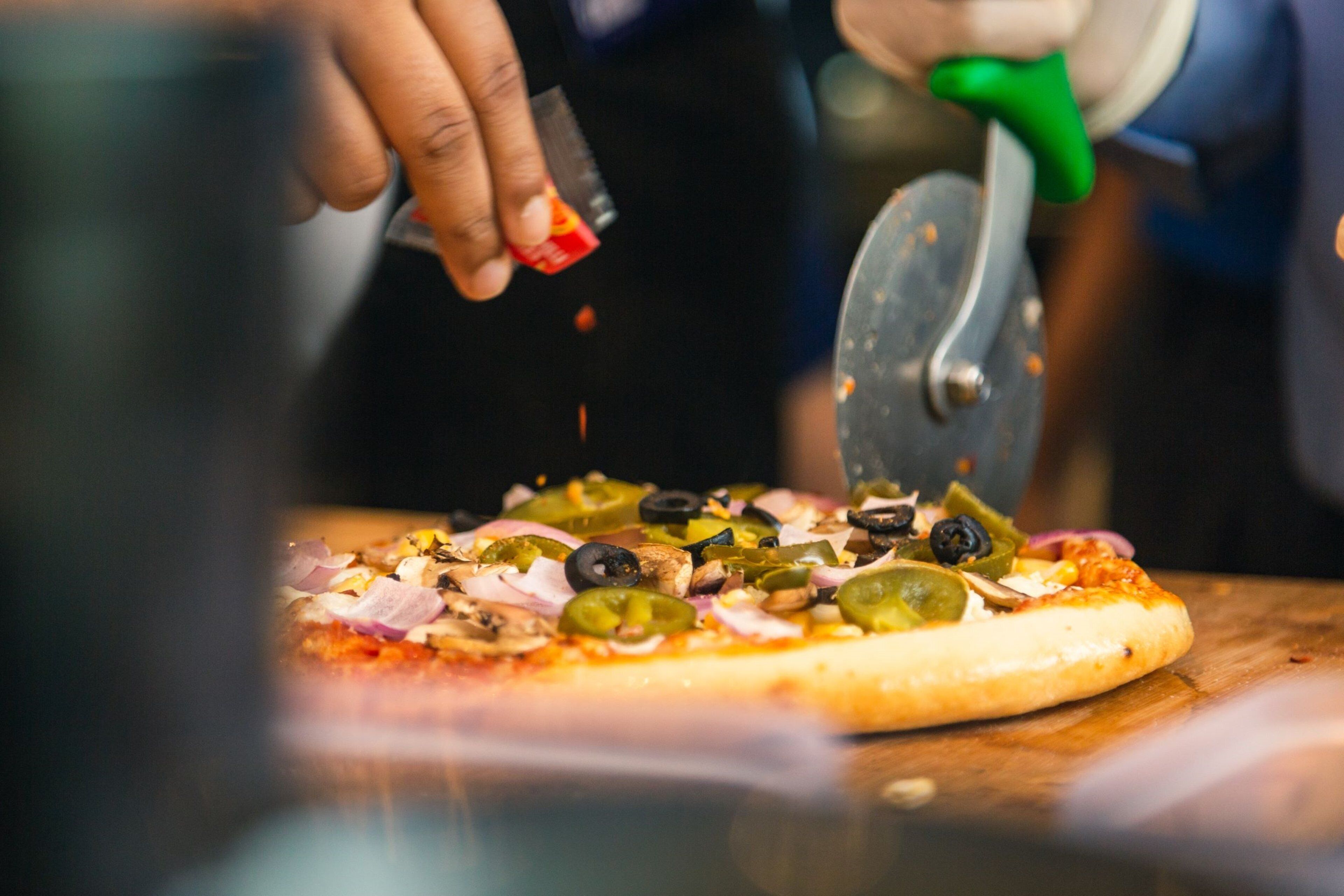Eine Pizza wird mit einem Pizzaschneider mit grünem Griff auf einer Holzunterlage geschnitten.