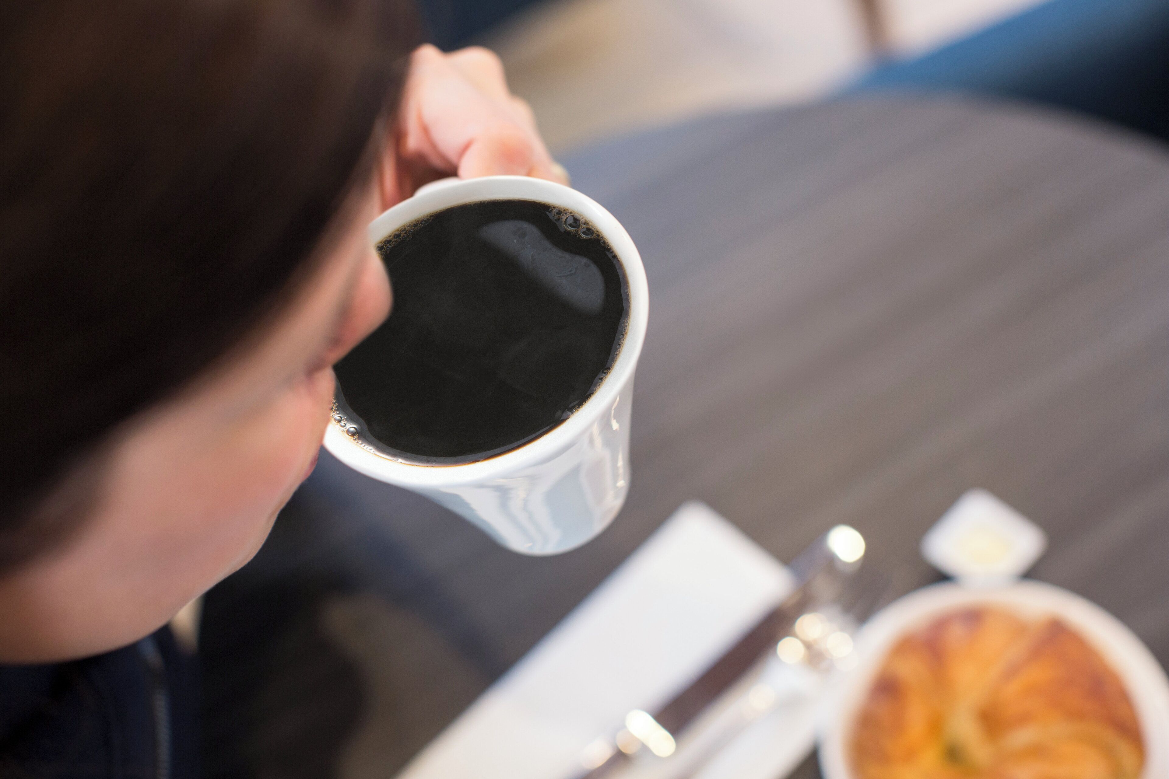 Una persona sosteniendo una taza de café, con un croissant y un plato pequeño sobre la mesa.