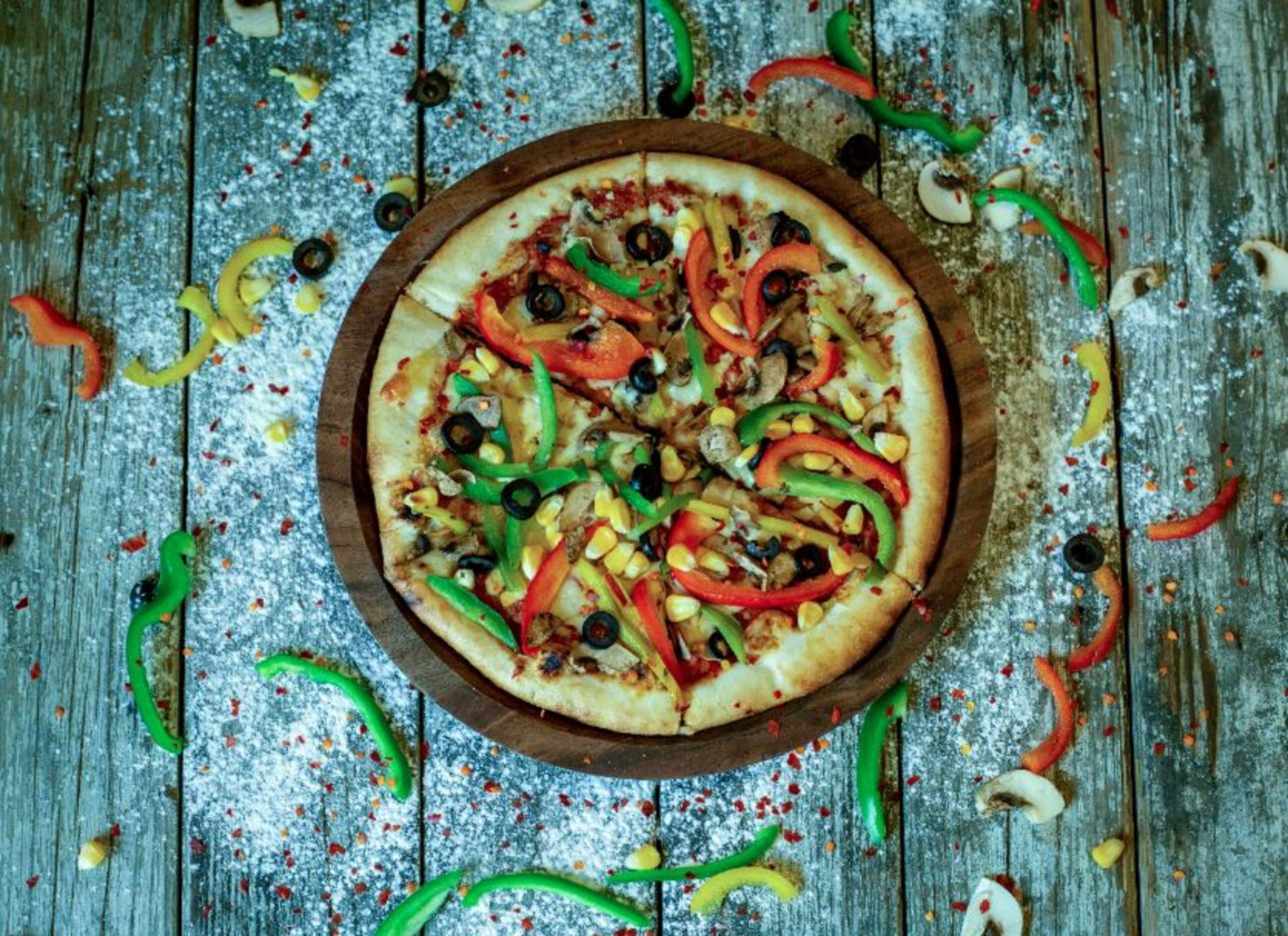 En pizza på en träbricka med olika sorters pålägg, omgiven av utspridda grönsaker på en träyta.