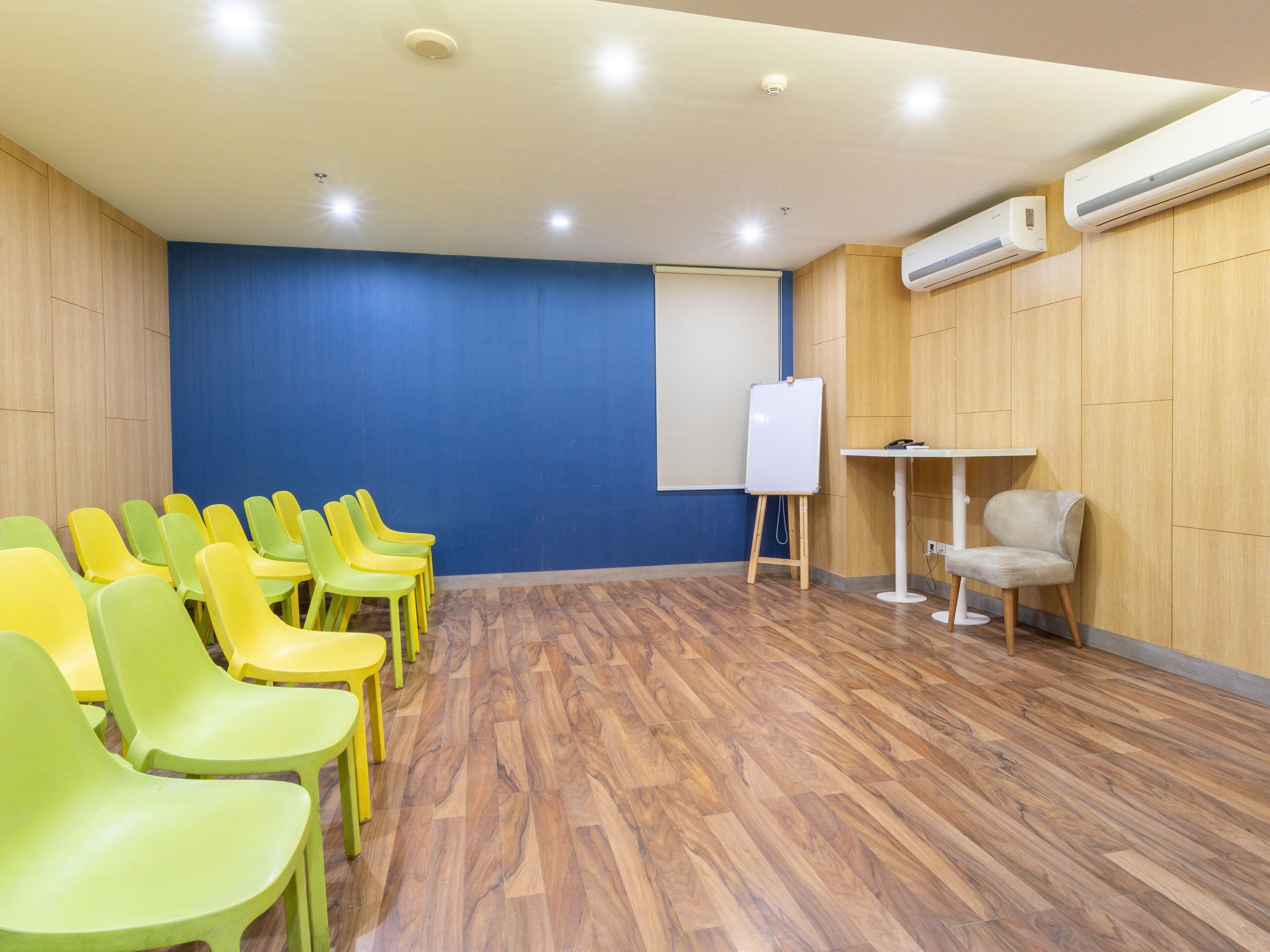 Une salle de conférence aux murs recouverts de panneaux en bois, avec un mur d’accent bleu et un tableau blanc. On y trouve des rangées de chaises jaunes et vertes, une table blanche et un appareil de climatisation fixé au mur.