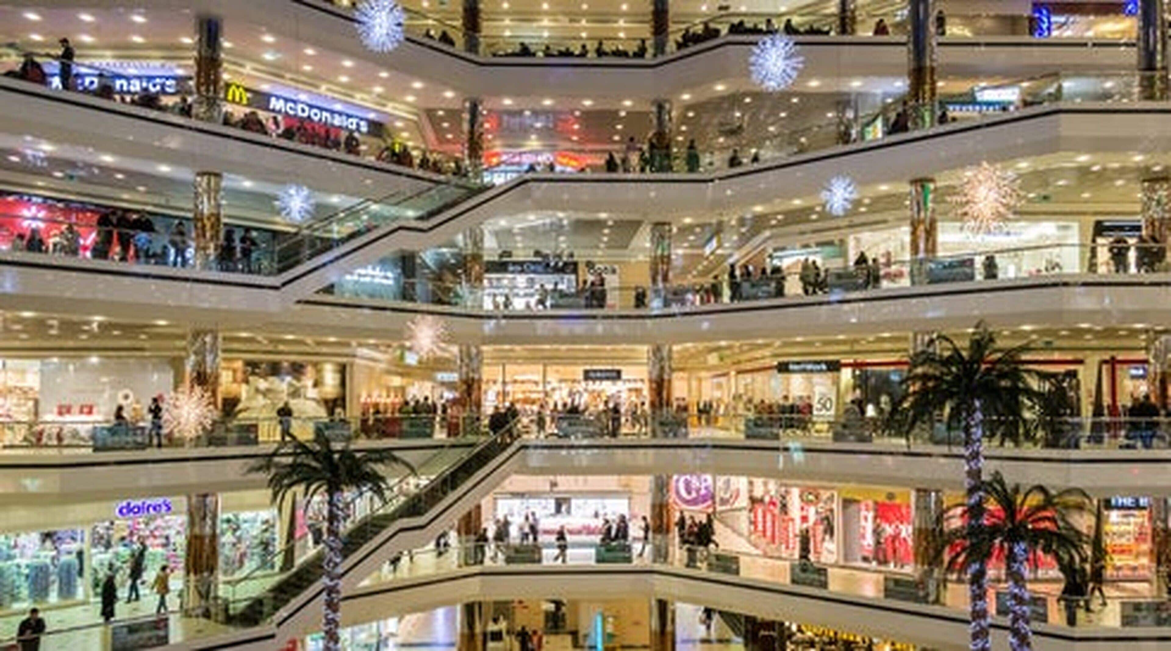 A multi-level shopping mall with escalators connecting floors, various retail stores, and decorative snow-like ornaments.