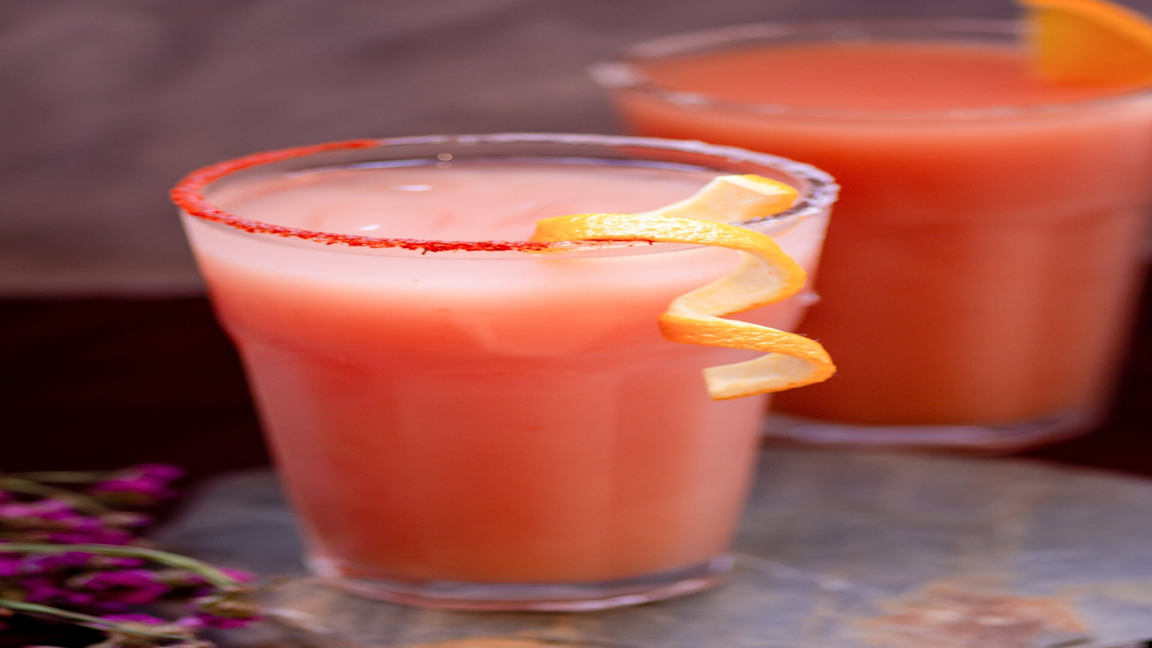 A tall glass with a pink beverage, garnished with an orange peel, on a wooden surface.