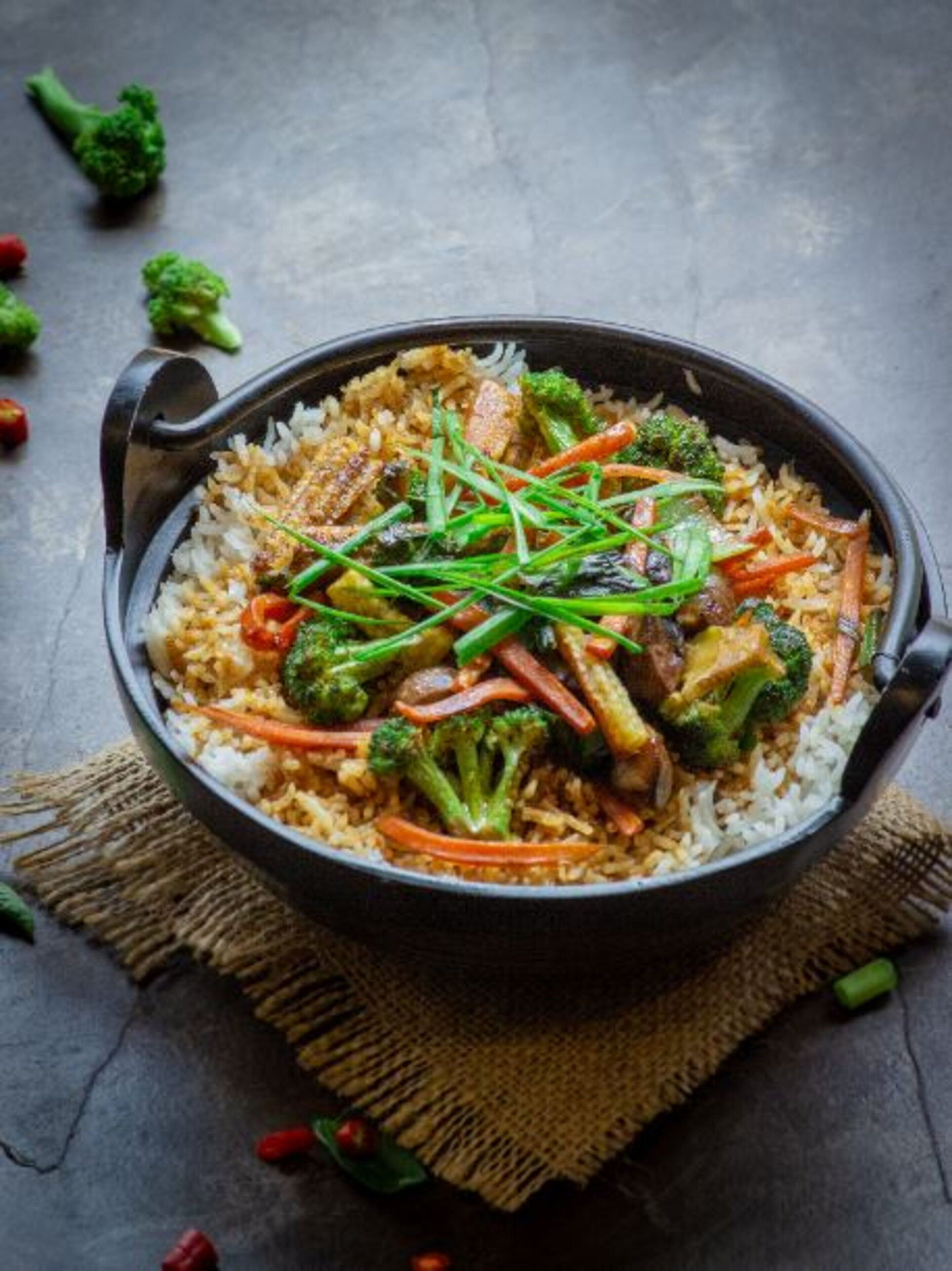 A bowl of fried rice with vegetables and meat, placed on a dark surface with a rustic burlap cloth underneath.