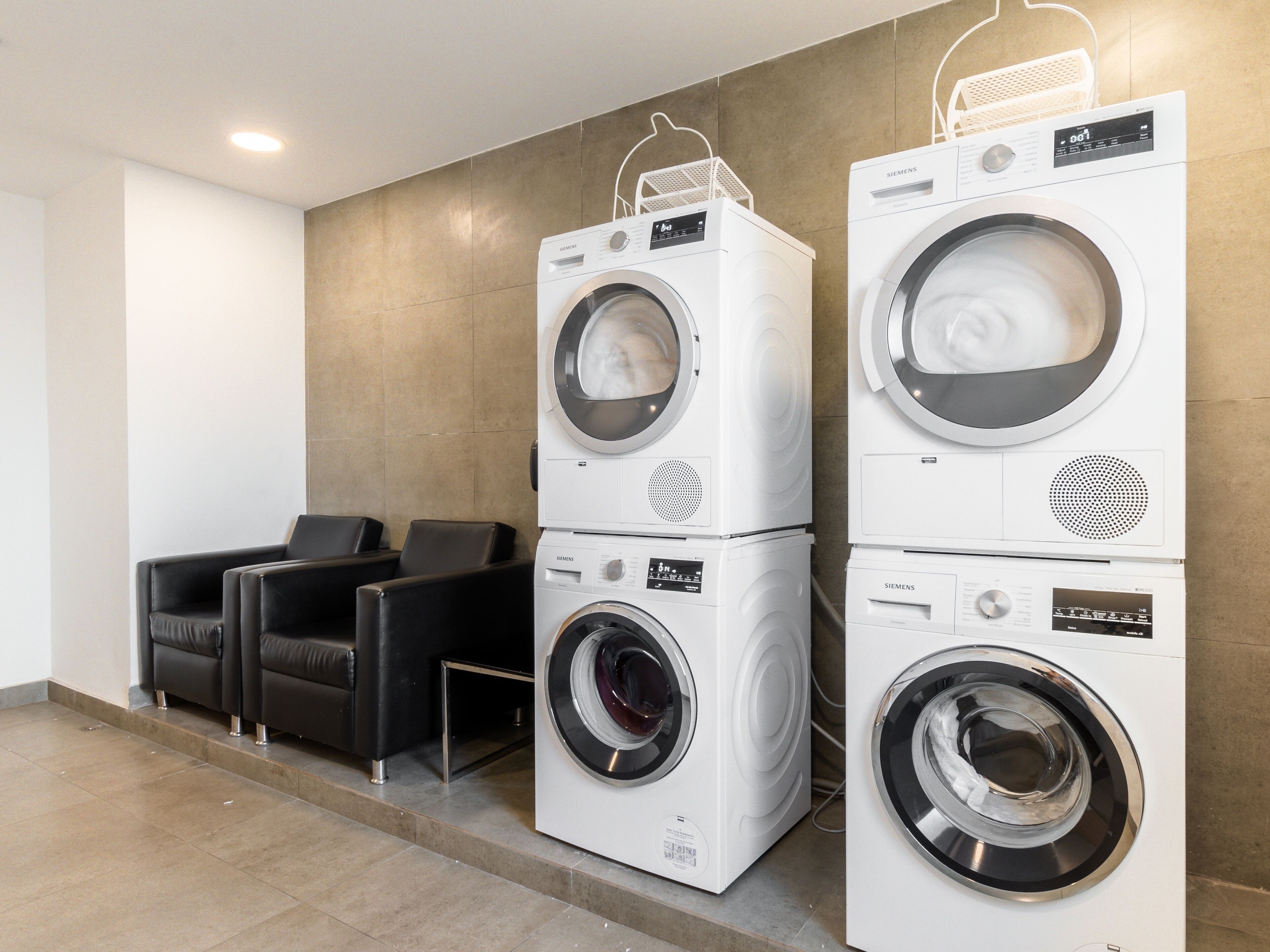 A laundry area with two stacked front-loading washing machines, a seating area with black chairs, and a tiled floor.