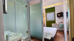 A modern hotel room with a glass shower enclosure, a white sink, a bed with white linens, a desk with a chair, and a mounted TV. - Hotel Caspia Pro Greater Noida (Greater Noida)
