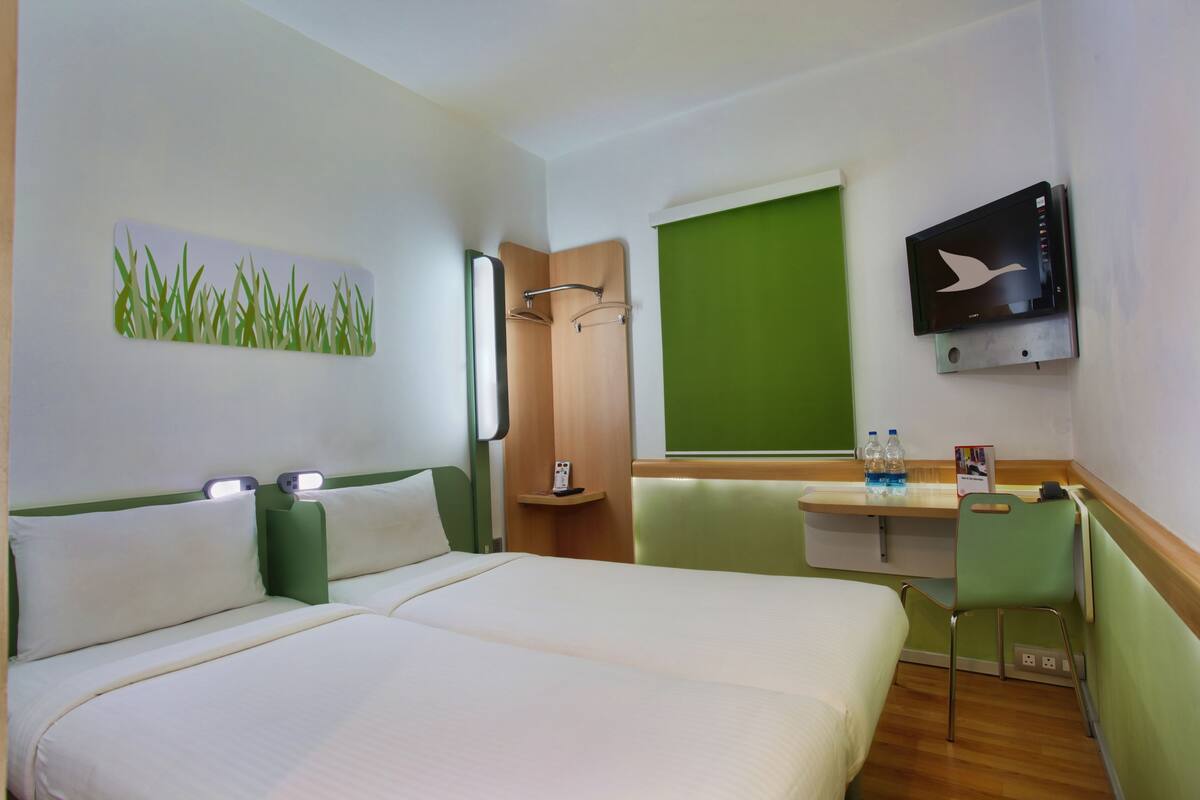 A hotel room with two beds, a desk, a chair, a television, and a green wall.
