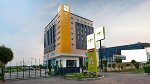 A hotel with a yellow and white facade, a signboard with a logo, and a parking area with a green fence. - Hotel Caspia Pro Greater Noida (Greater Noida)