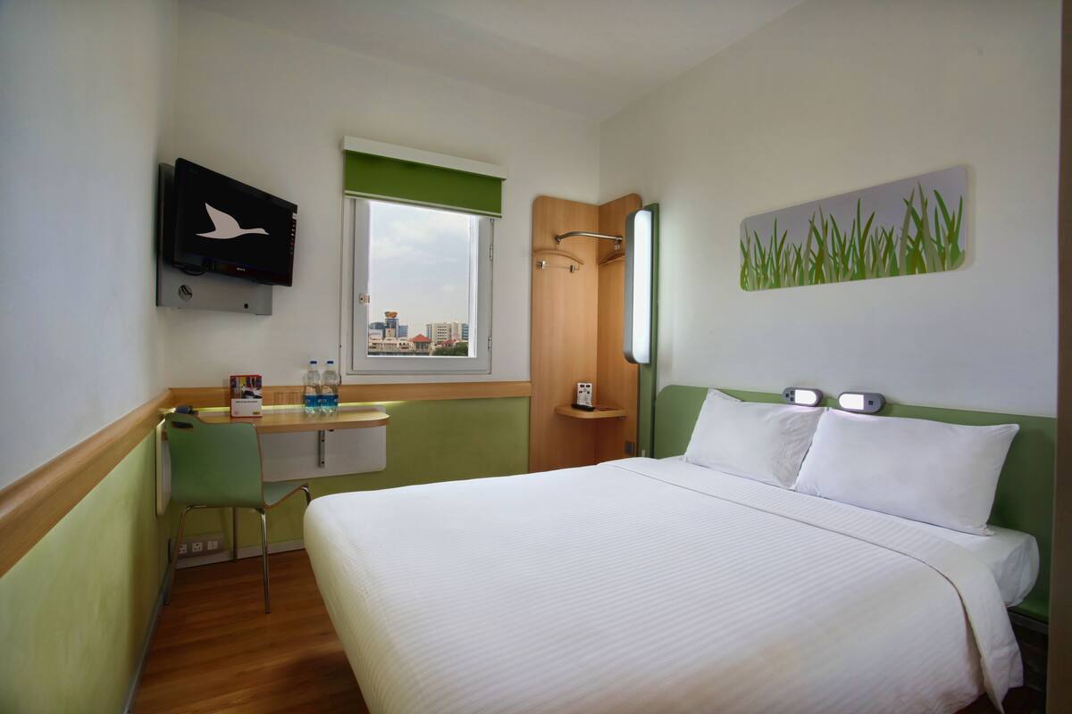 A hotel room with a bed, a desk with a chair, a television, a window, and a painting on the wall.