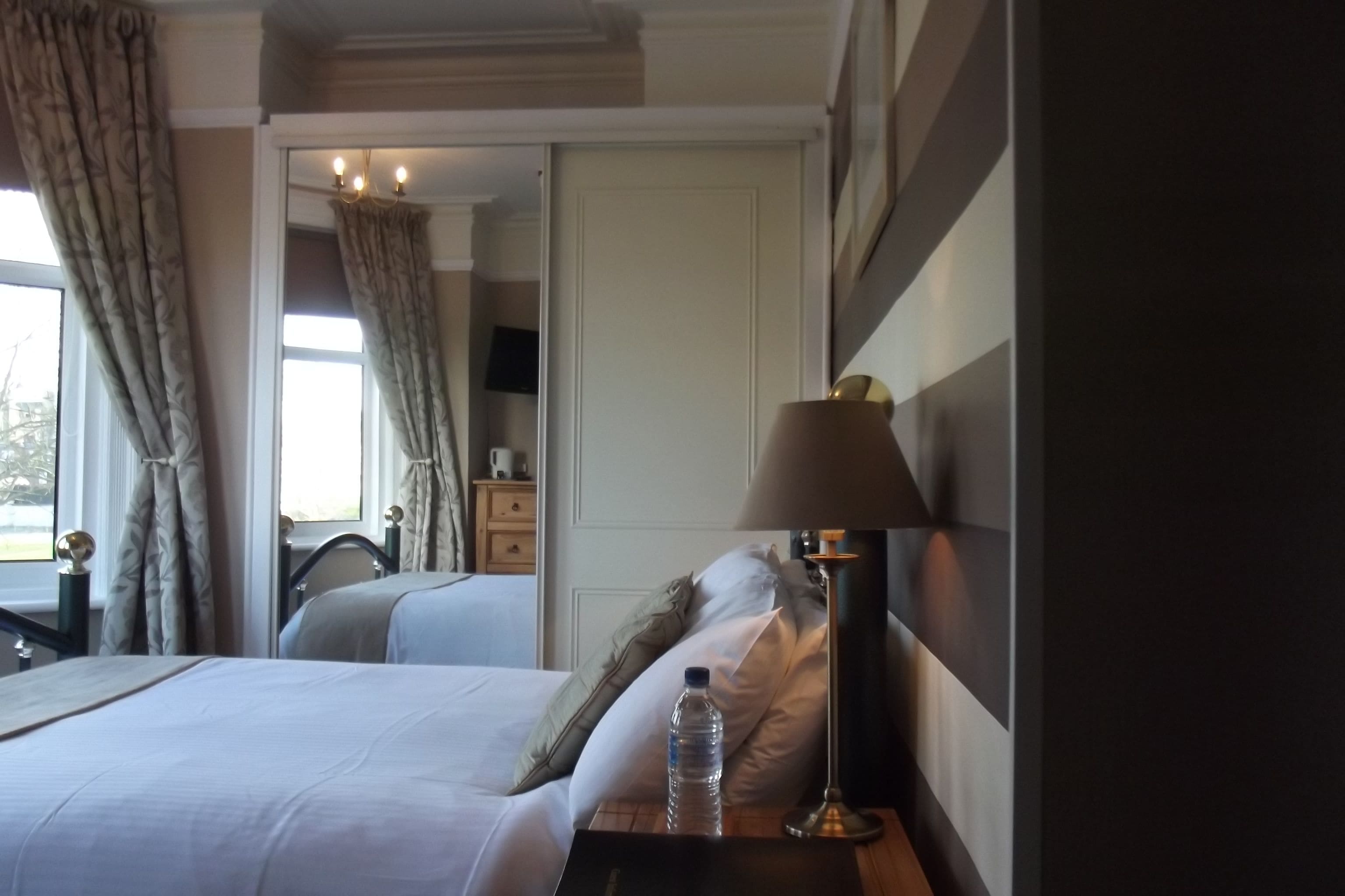 A hotel room with a bed, a nightstand with a lamp, a water bottle, and a mirror.