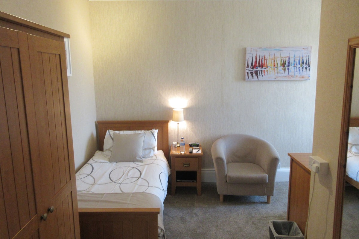 A hotel room with a bed, a chair, a nightstand with a lamp, a painting on the wall, and a trash bin.