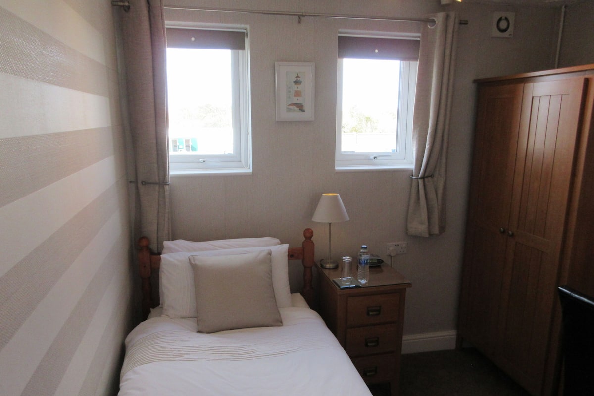 A bedroom with a bed, a nightstand, a wooden wardrobe, two windows, and a wall-mounted picture.
