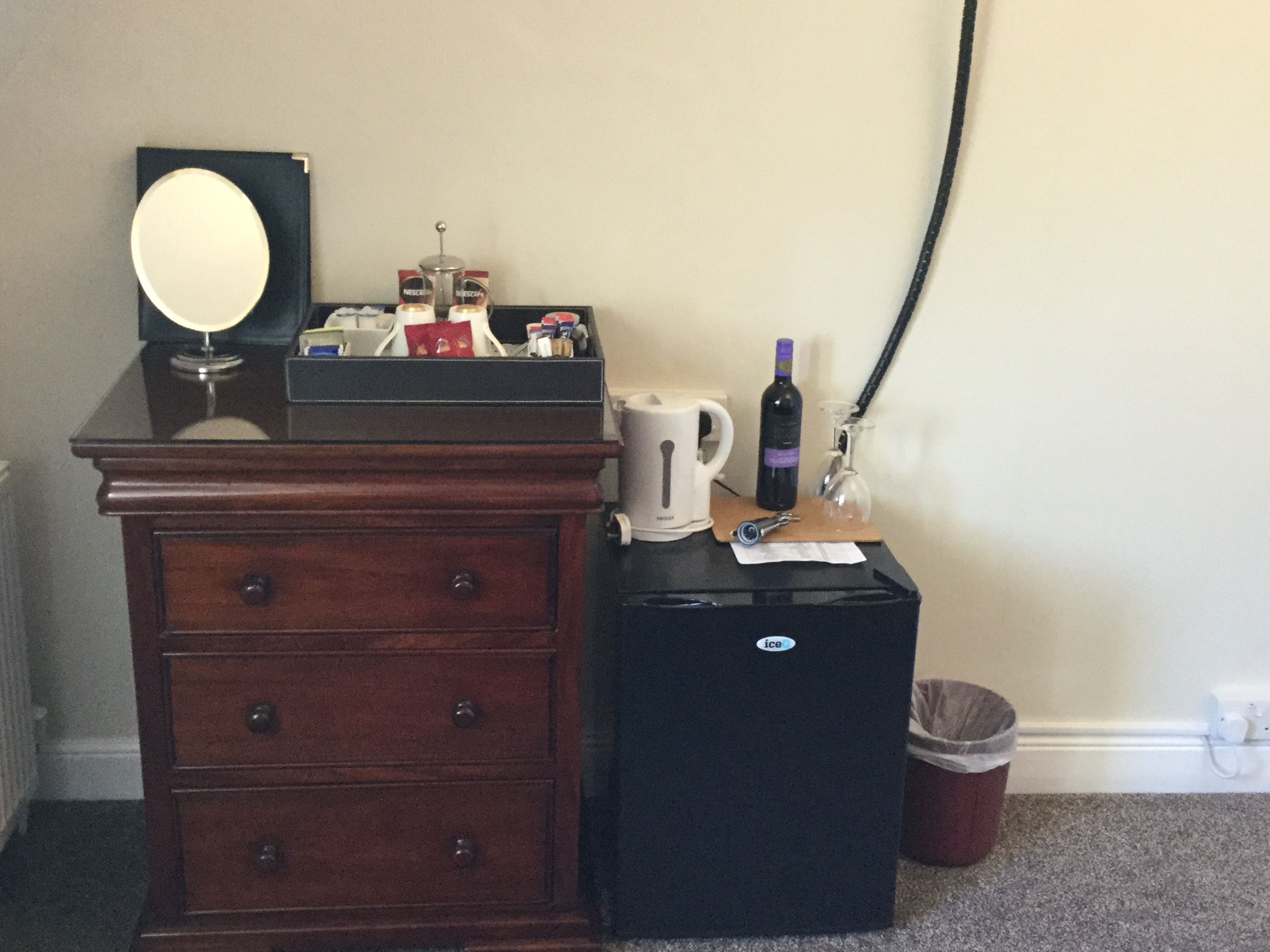 A wooden dresser with drawers, a small black refrigerator, a white kettle, a bottle of wine, a glass, and a trash can.