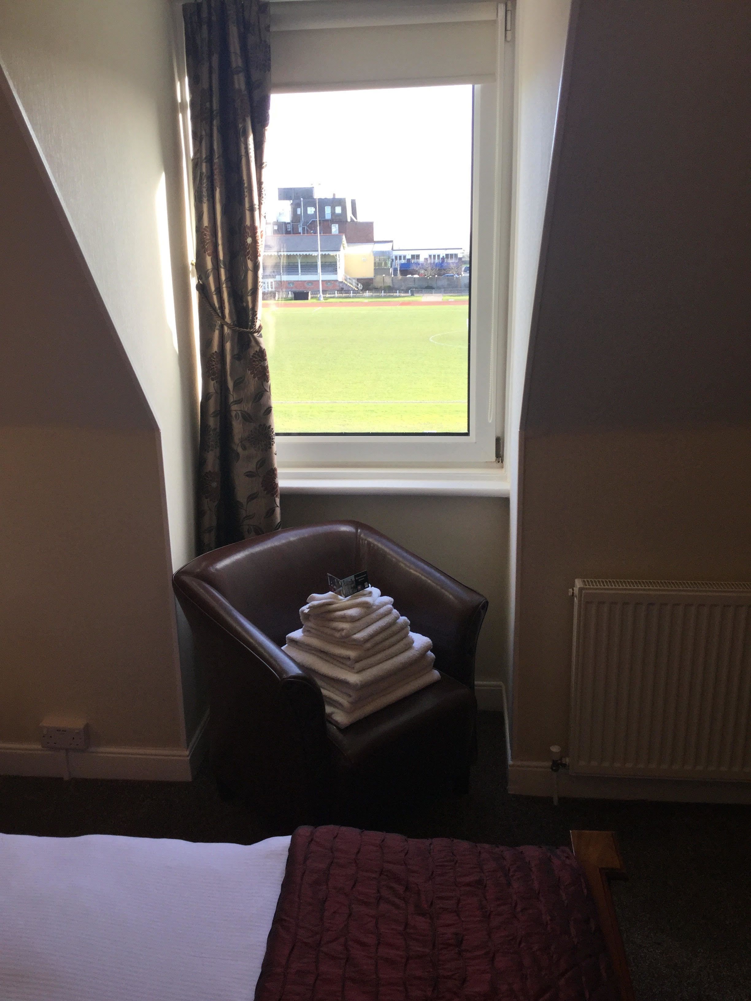 A hotel room with a bed, a chair with folded towels, a window with a view of a sports field, and a radiator.