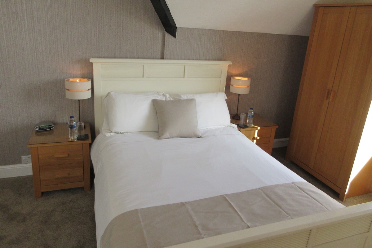 A neatly made bed with white linens, two bedside lamps, a water bottle, and a digital clock on a wooden nightstand.