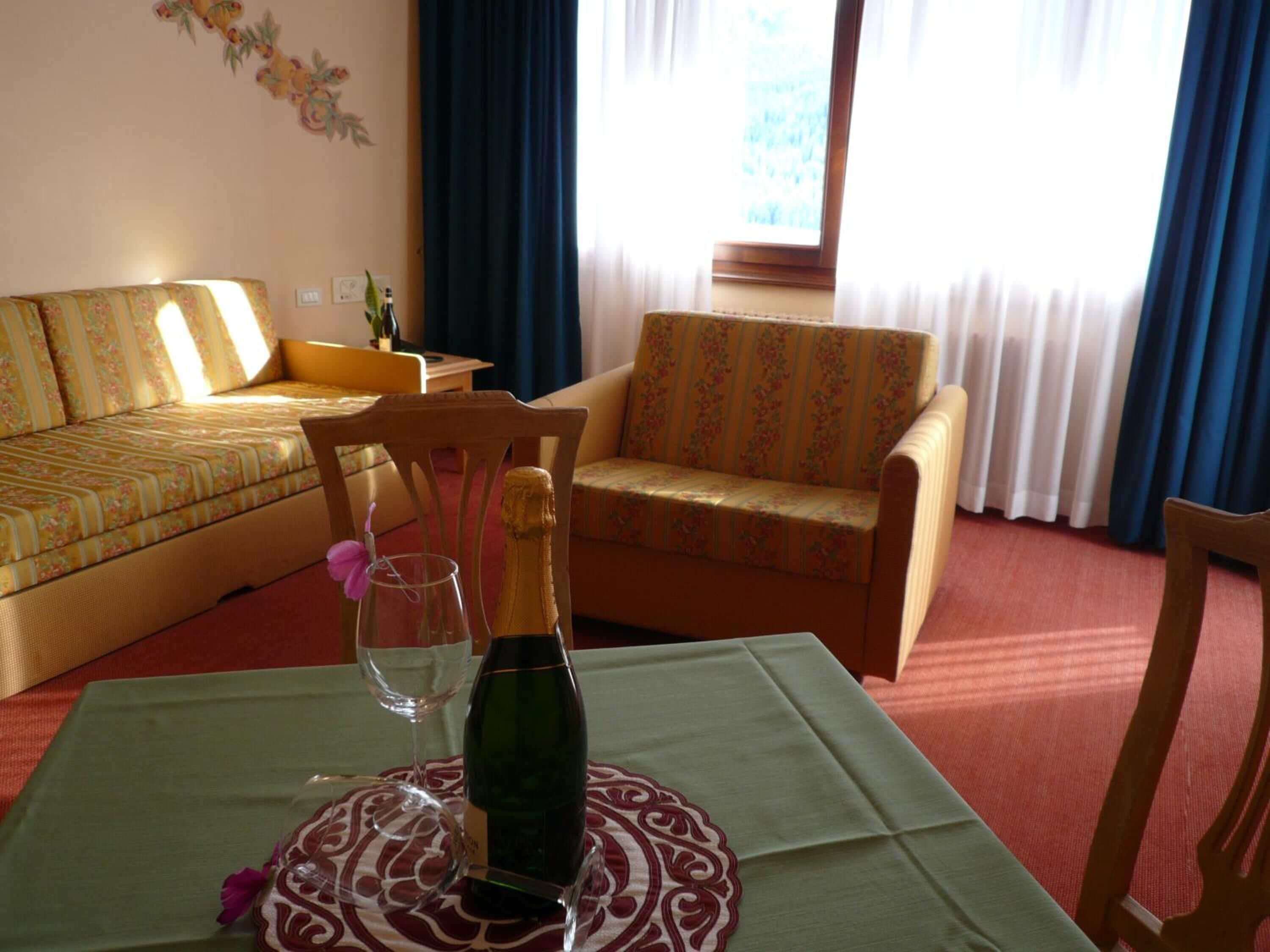 A living room with a sofa, a chair, a table with a bottle of wine and a glass, and a window with curtains.