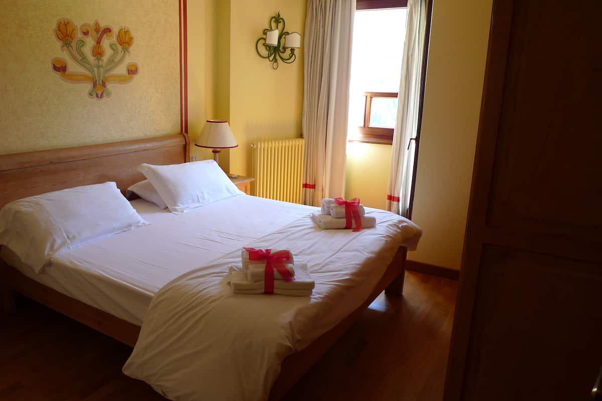 A neatly made bed with white linens and towels, a wooden headboard, a wall-mounted lamp, and a window with curtains.