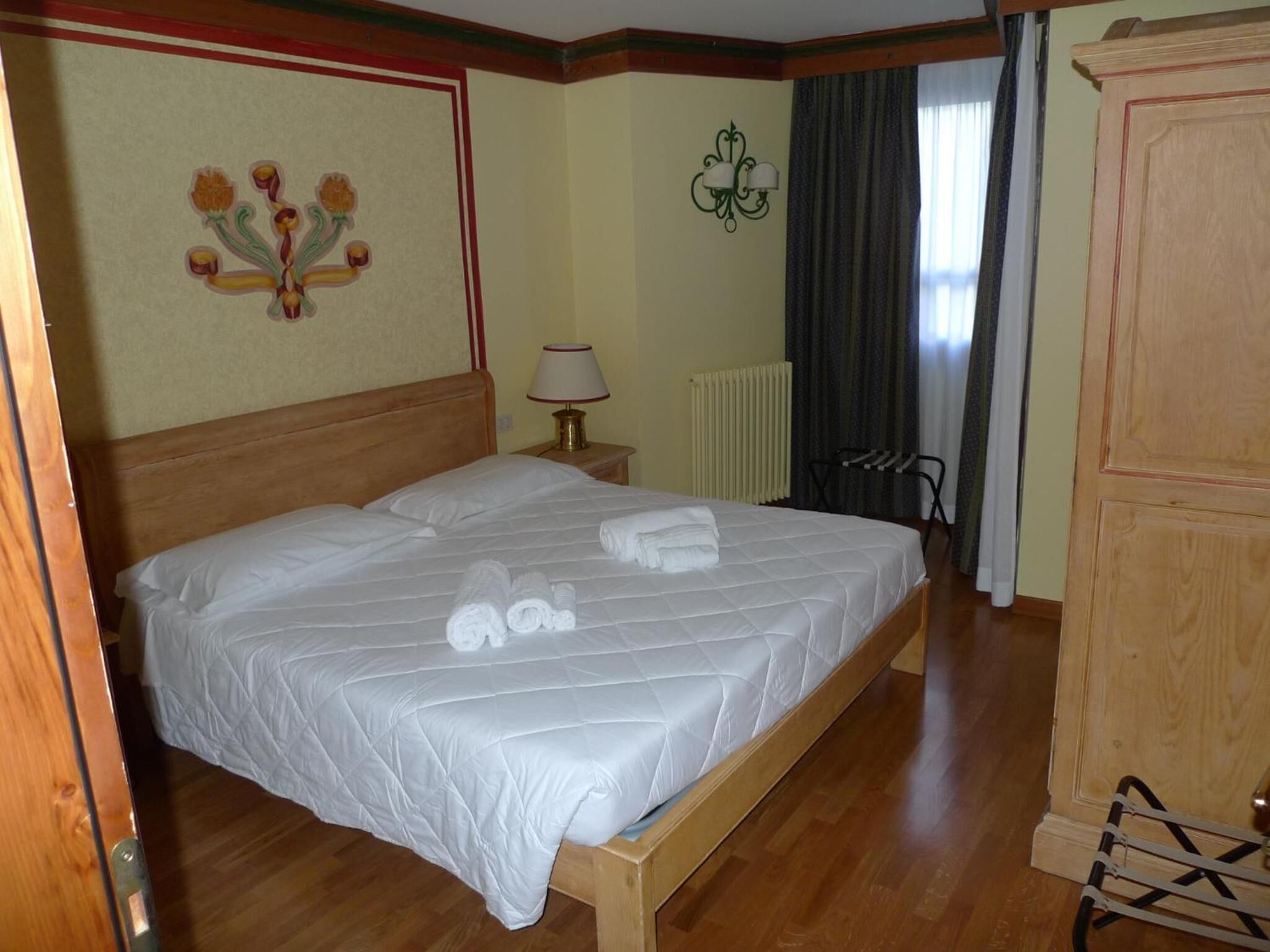 Una cama bien hecha con sábanas y toallas blancas, cabecera de madera, lámpara de pared y radiador.
