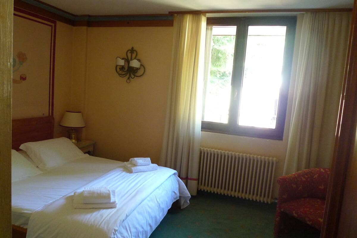 A hotel room with a bed, a nightstand, a lamp, a radiator, and a window with curtains.