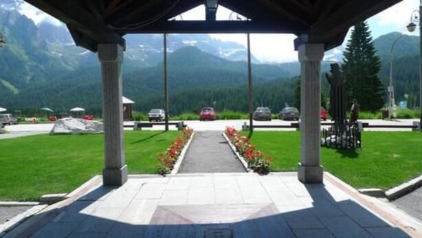 A covered walkway leading to a parking area with cars and tents, surrounded by mountains and greenery. - Hotel & Residence Catturani (Pinzolo)