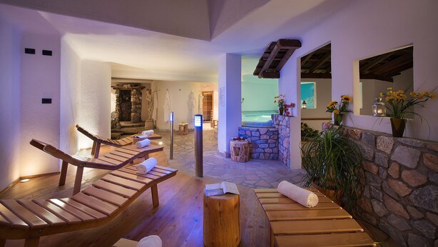 A spa area with wooden loungers, a stone wall, and a view of the ocean. - Hotel Cristallo (Moena)